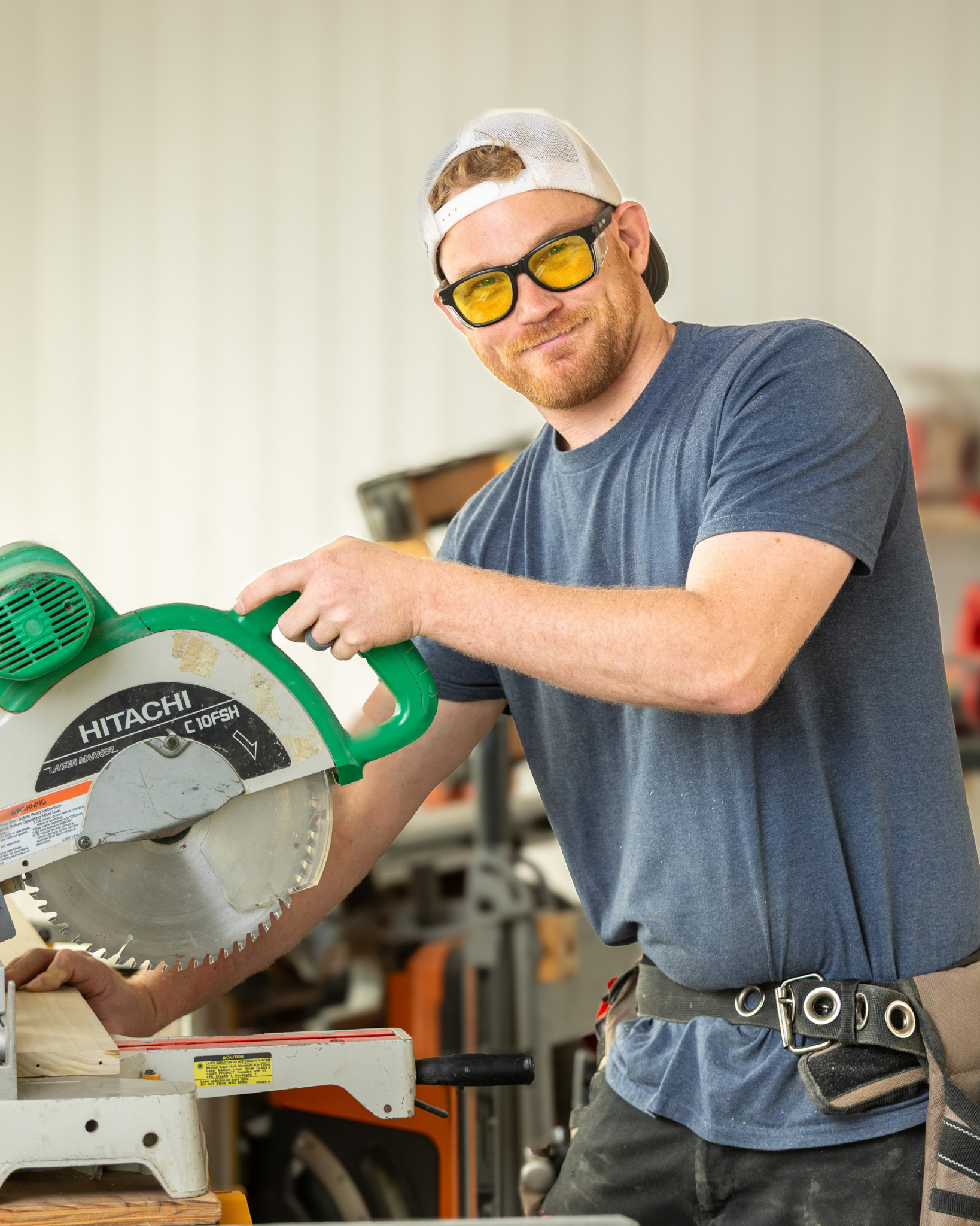 Trent Wilson is a young man wearing a white backwards cap, yellow glasses, and a dark blue t-shirt using a green Hitachi circular saw in a workshop.