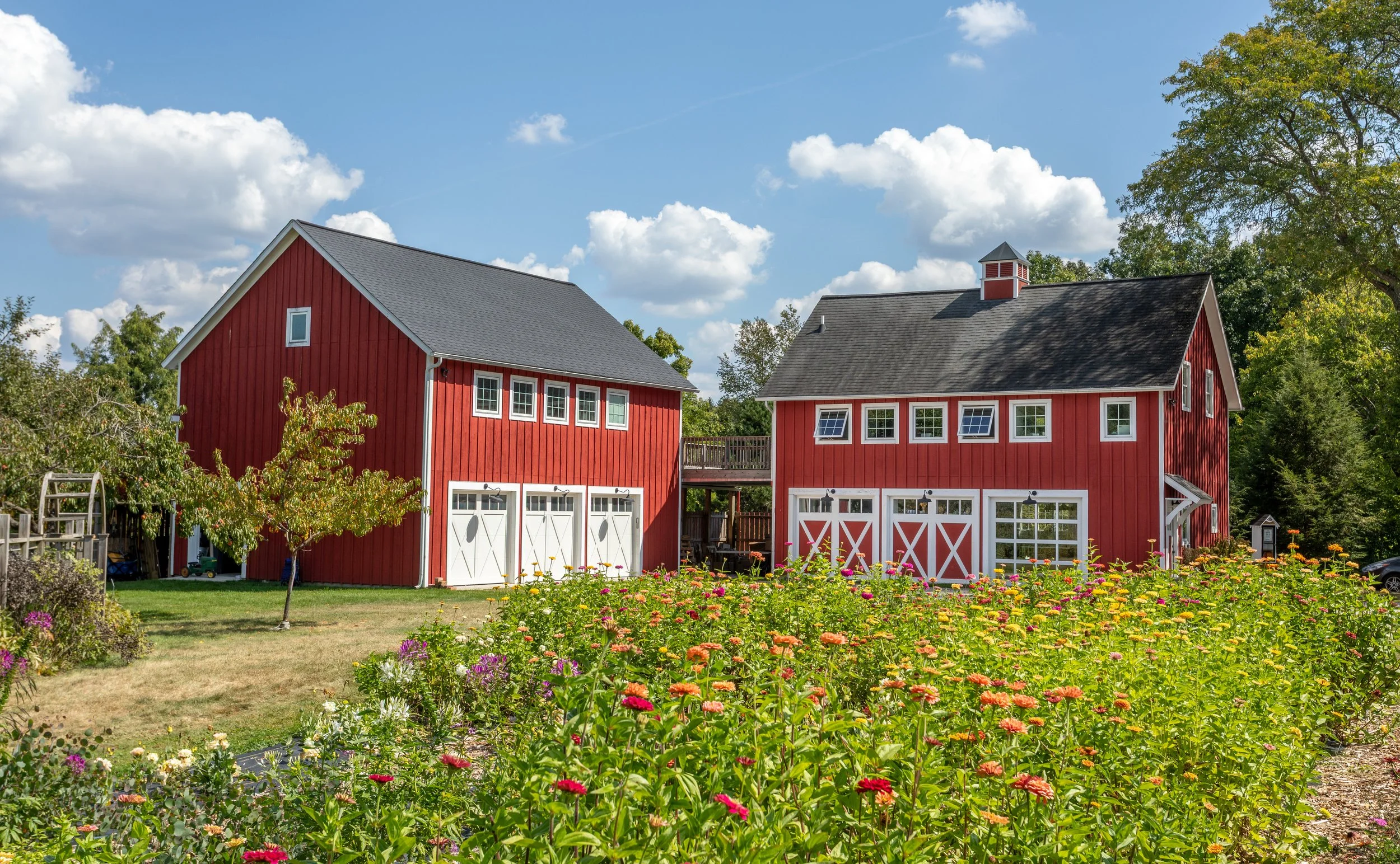 Two red barns at artisan business Little Flower Soap Co., with white doors and trim, surrounded by a garden with colorful flowers and green trees under a blue sky with white clouds.