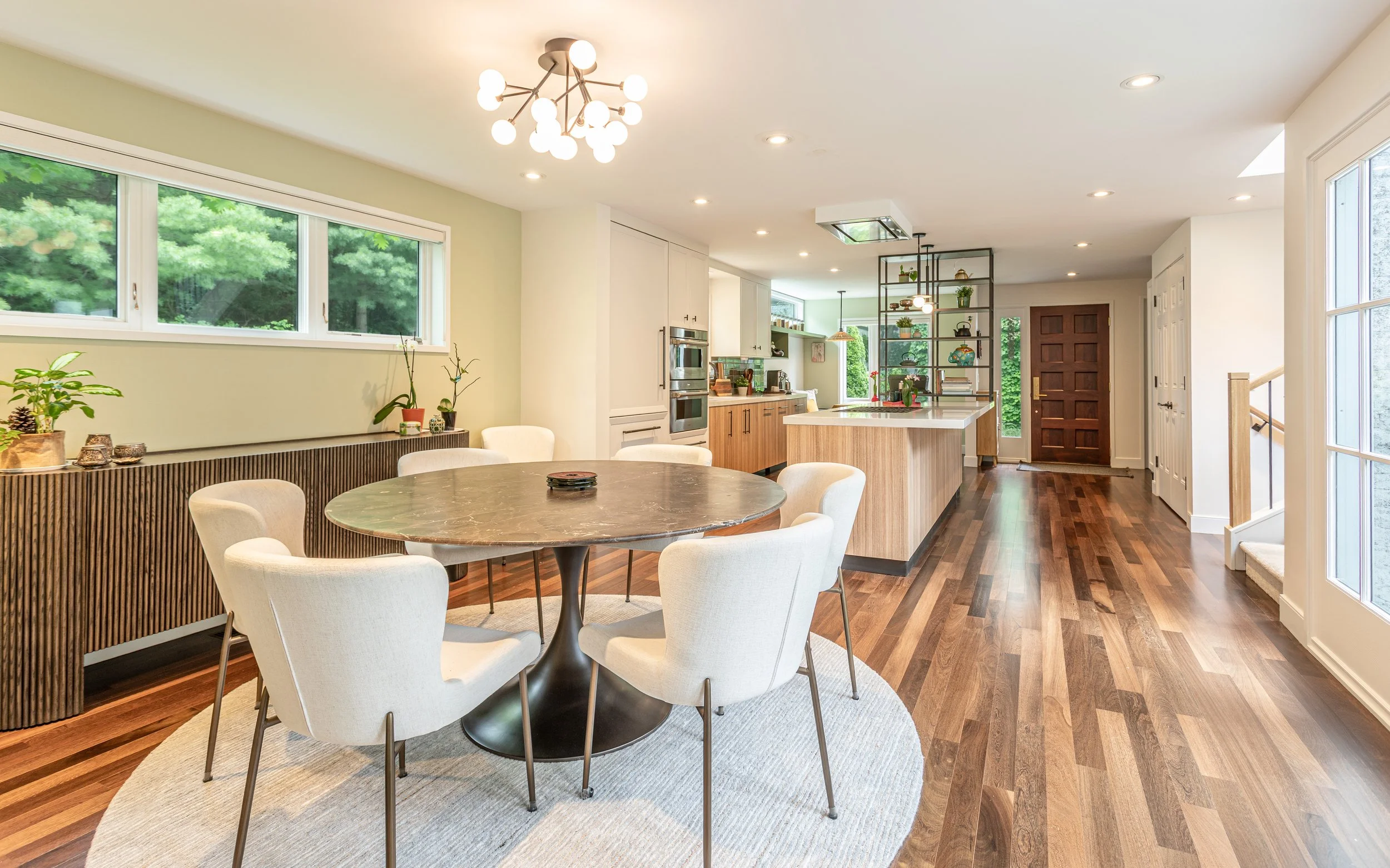 Open-concept kitchen and dining area with wooden flooring, a round dining table with six white chairs, large window with green trees outside, modern chandelier, kitchen island with shelving, and built-in appliances.