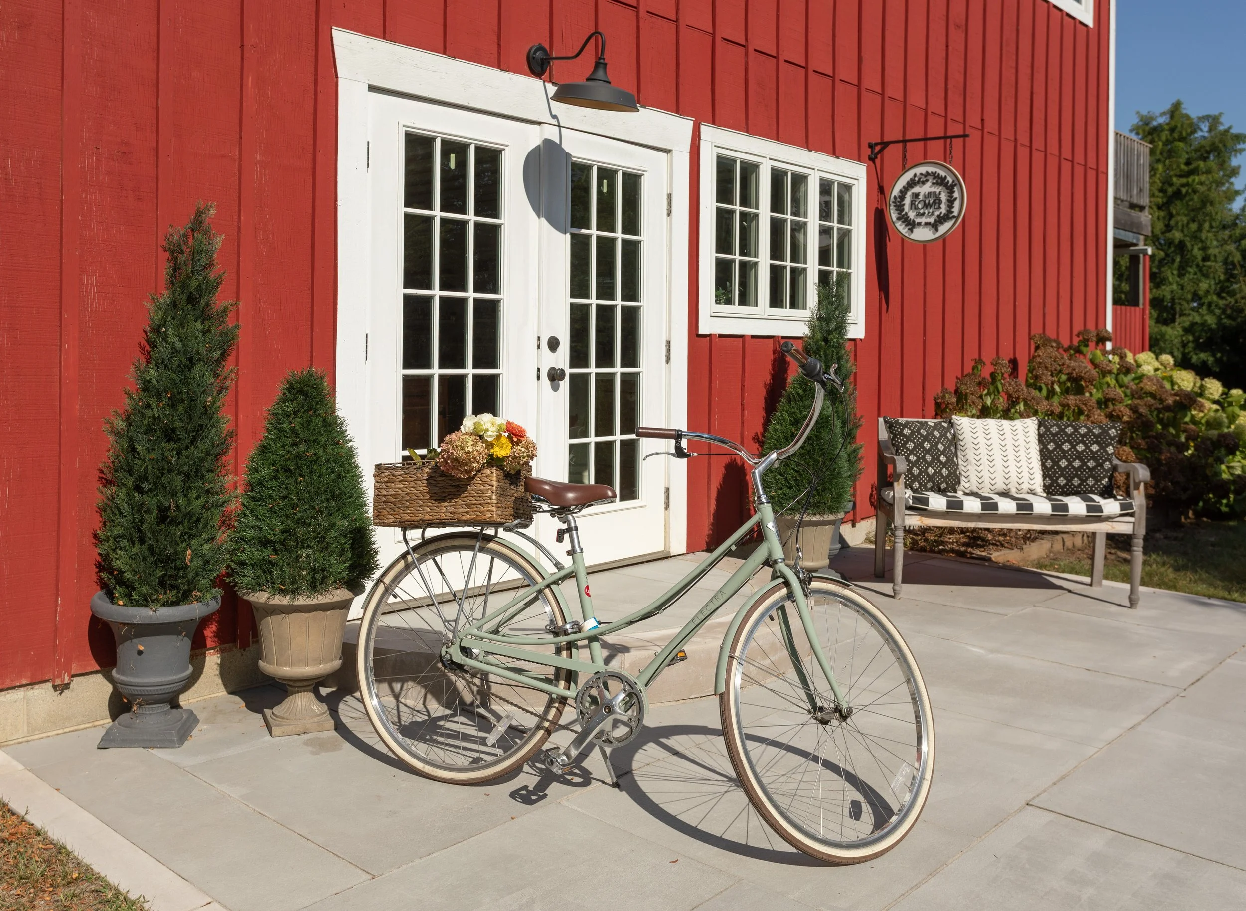 Red barn for Little Flower Soap Co. with white double doors, potted trees, a bicycle with a basket of flowers, and a bench with patterned cushions outside.