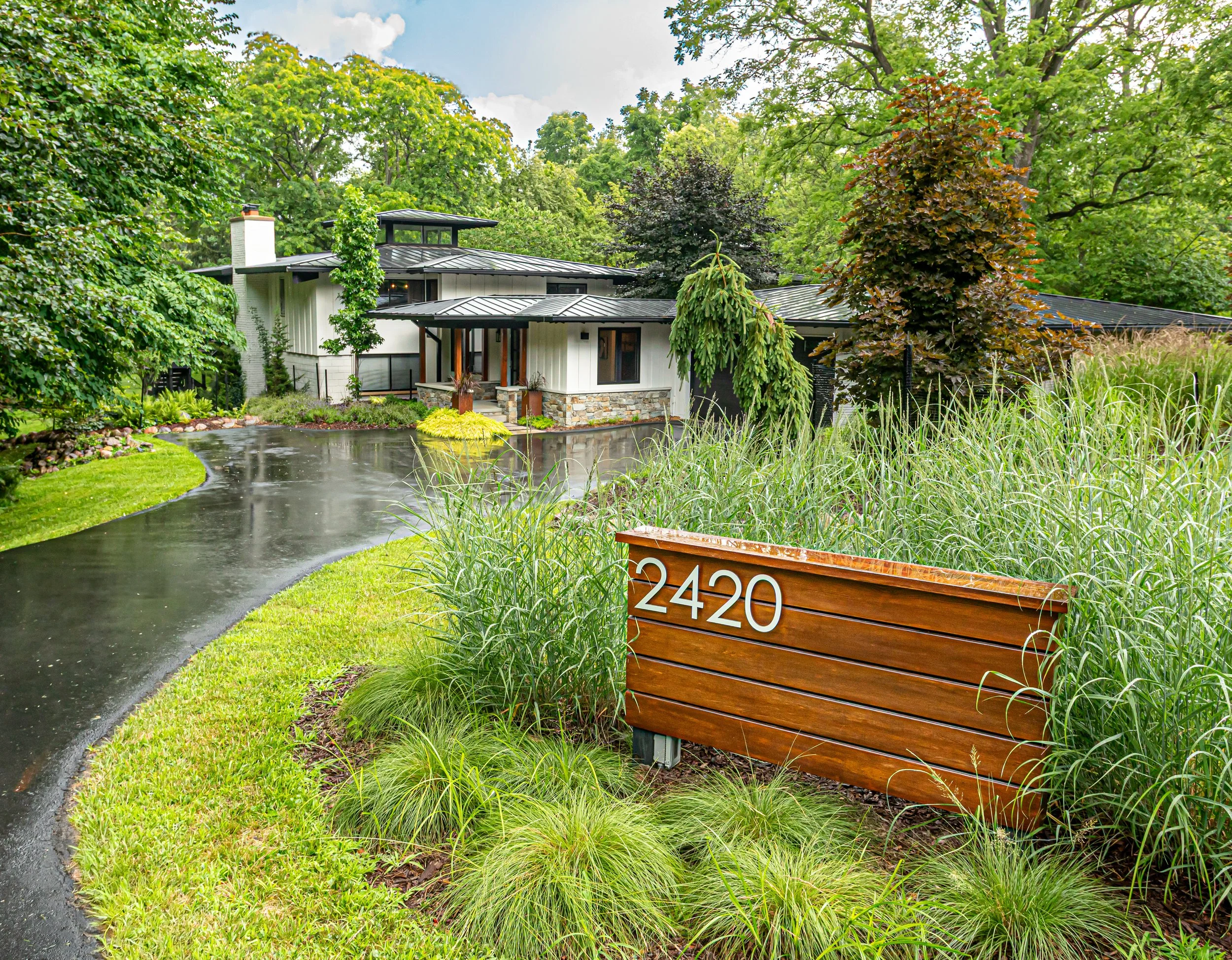 Modern custom luxury home with a curved driveway, lush greenery, and a wooden sign with the number 2420 in the front yard.