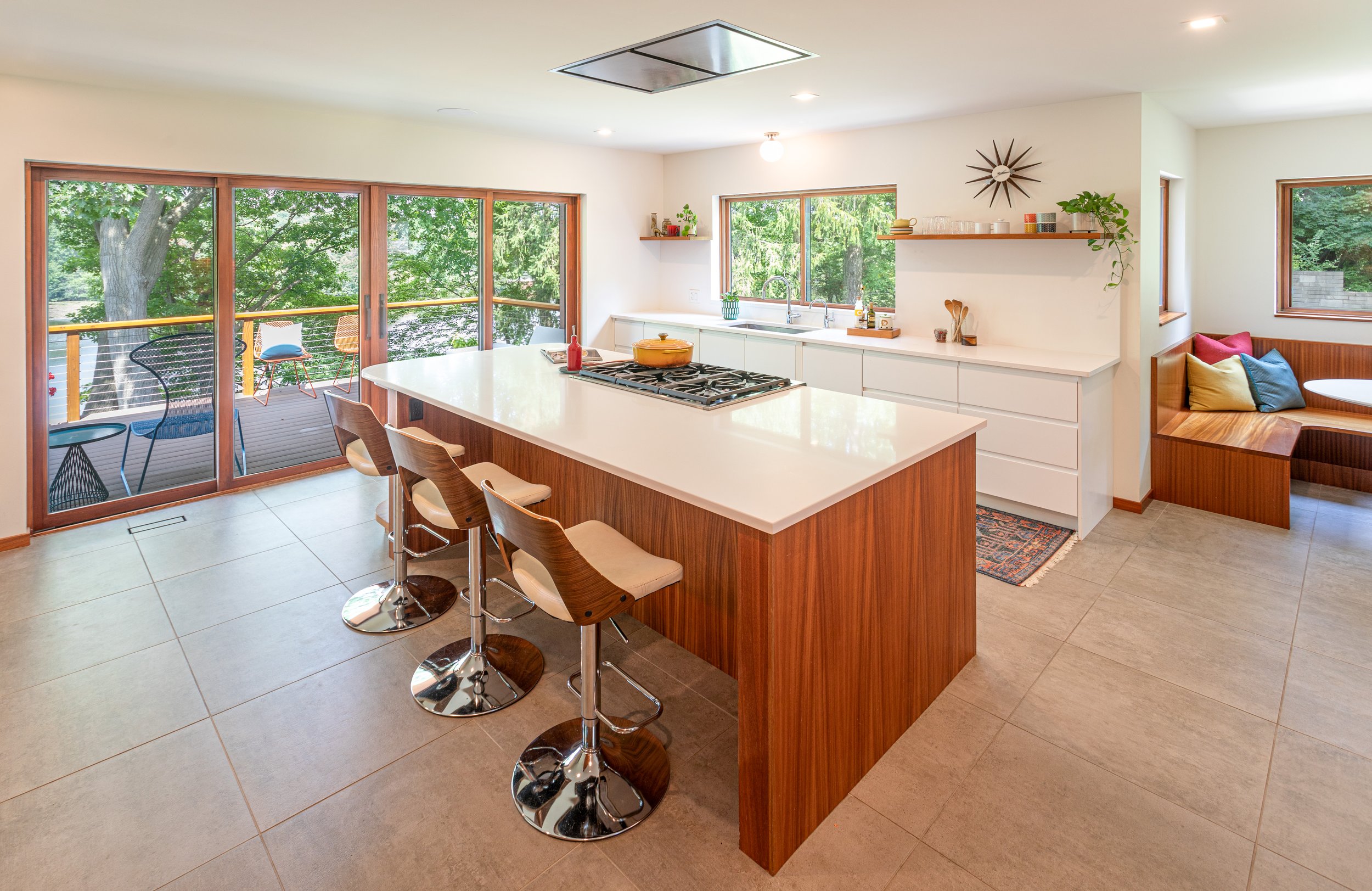 Modern kitchen with white countertops, wooden accents, island with three bar stools, large sliding glass door leading to a balcony with outdoor seating, and large windows with views of green trees.