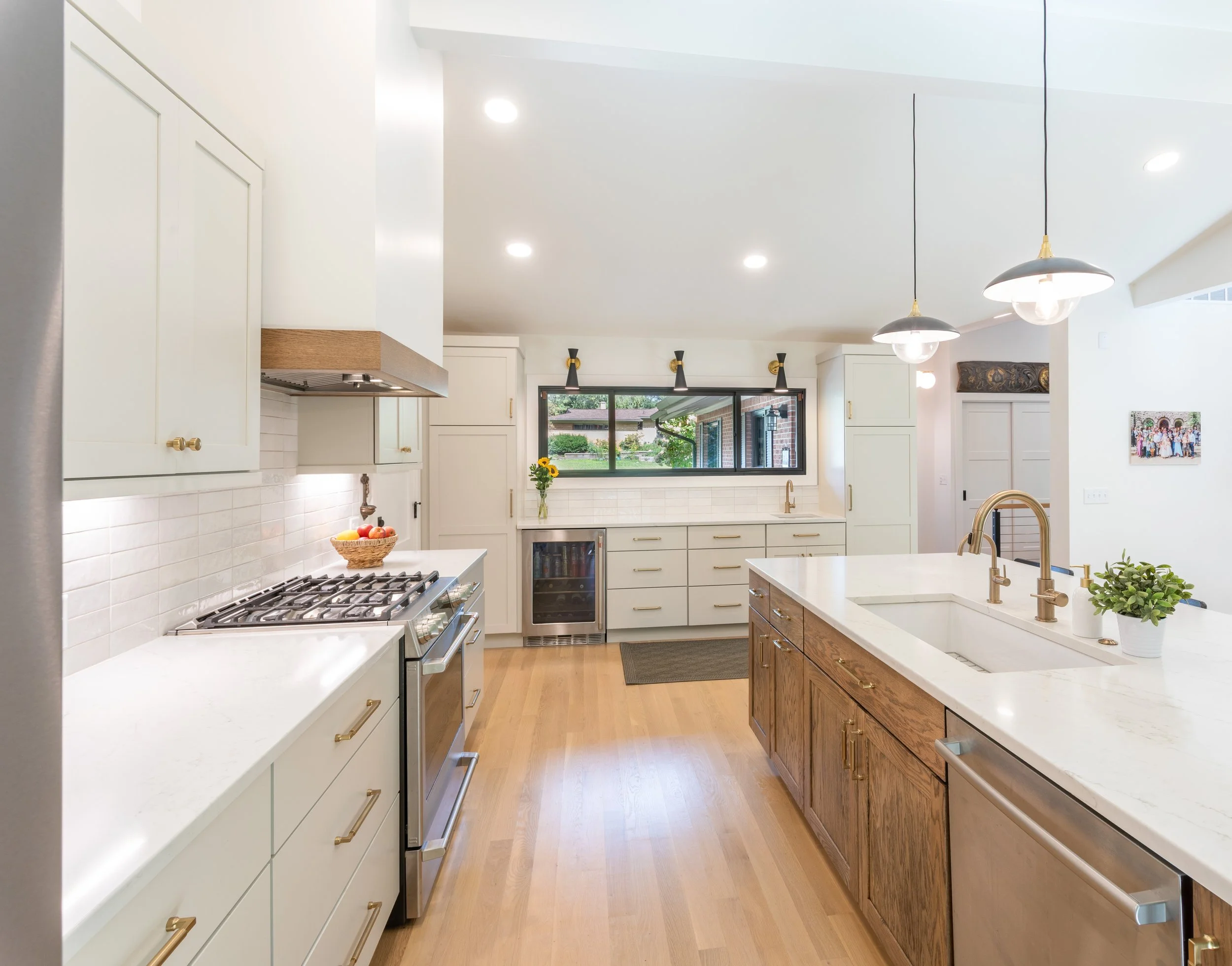 Modern kitchen with white and wooden cabinets, a large white marble island with a sink, brass fixtures, pendant and recessed lighting, a window with outdoor view, and hardwood flooring.