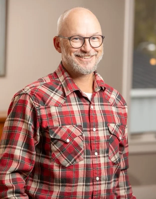 Shawn Lenhardt is a smiling middle-aged man with glasses and a beard wearing a red plaid shirt, standing indoors near a window.