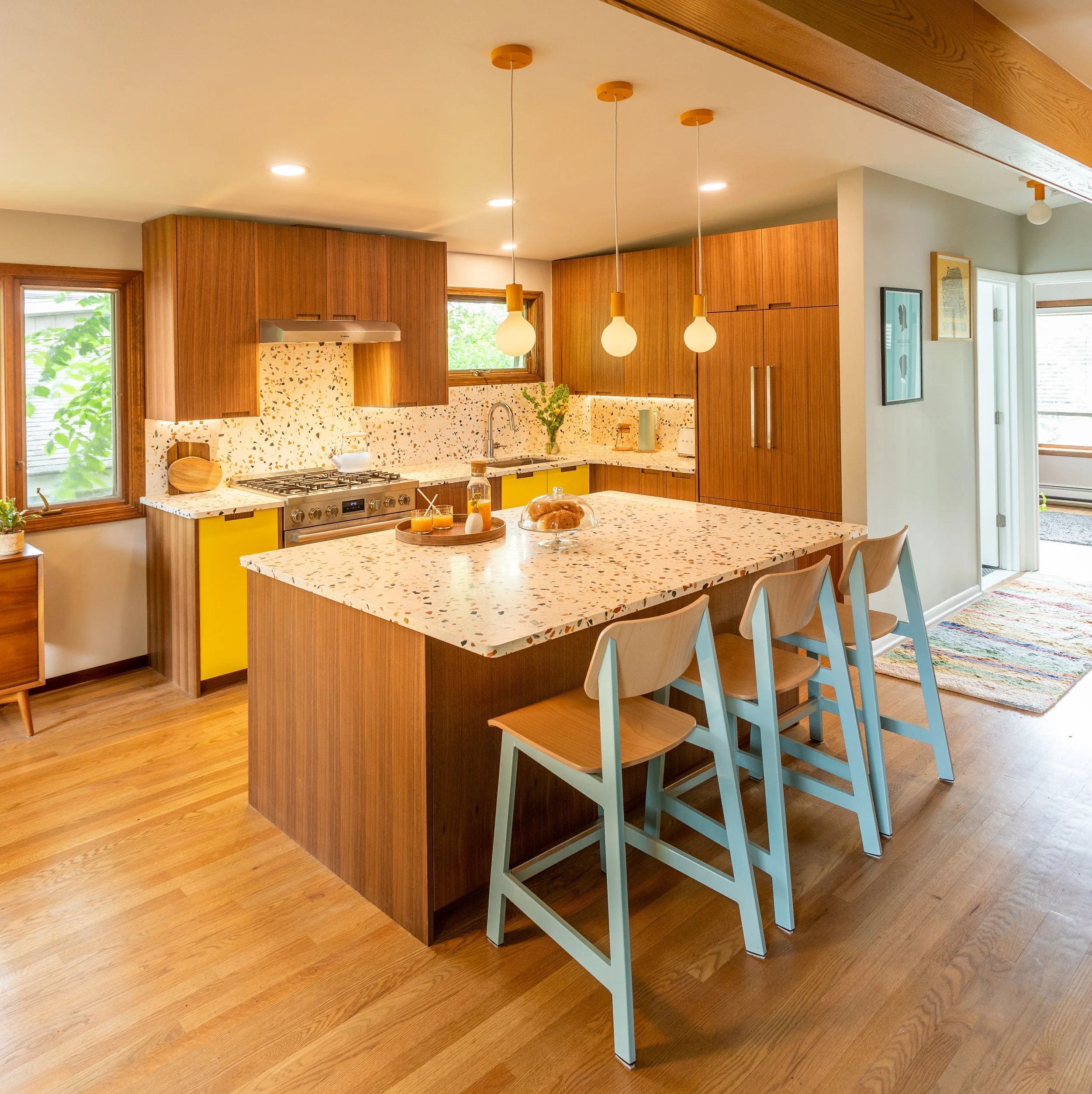 A modern kitchen with wooden cabinets, a speckled white countertop, a kitchen island with a matching speckled surface, three bar stools with white legs and wooden seats, pendant lights hanging over the island, a built-in oven, a window with greenery 
