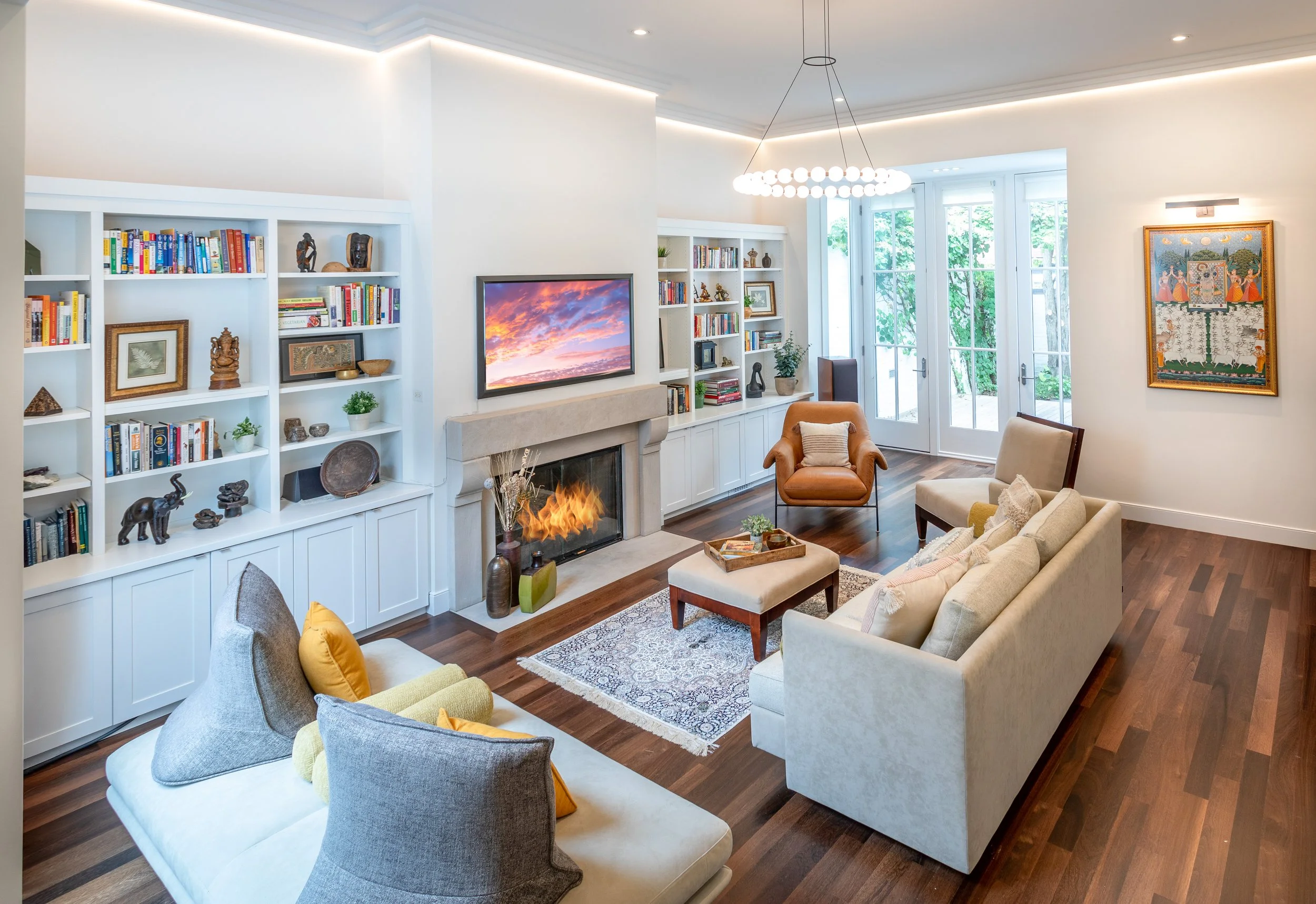 Living room with white walls, dark wood floors, built-in white shelves with books and decorative items, a fireplace, a wall-mounted TV above the fireplace, a glass door leading outside, an area rug, a beige sofa, a brown armchair, a beige chair, a small ottoman, and artwork on the walls.
