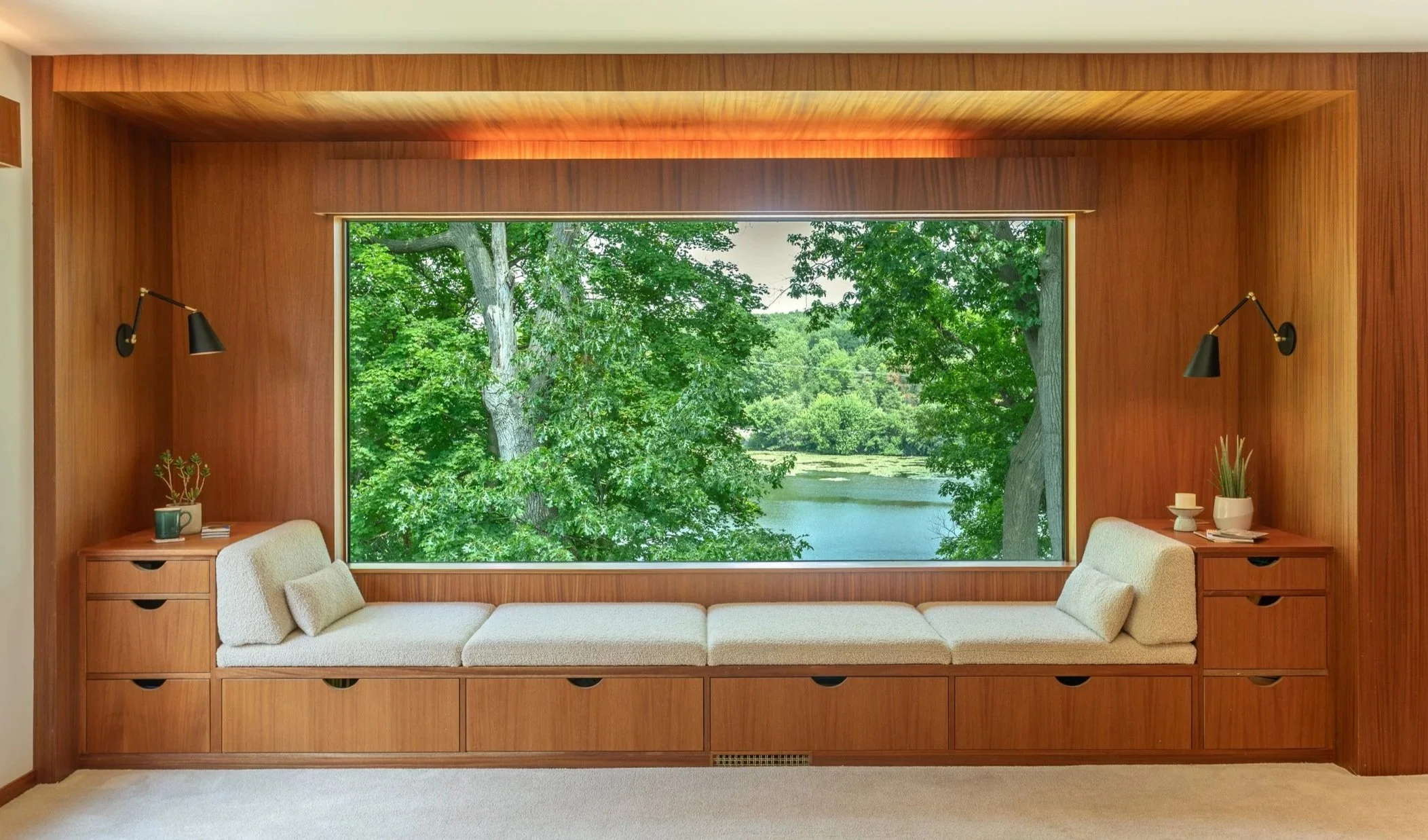 Cozy window nook with built-in cushioned seating in a mid-century modern home, surrounded by warm wood paneling, overlooking a lush green riverside view. This historic home was designed in 1938 by  Architect Ralph Stephens Gerganoff (R.S. Gerganoff).