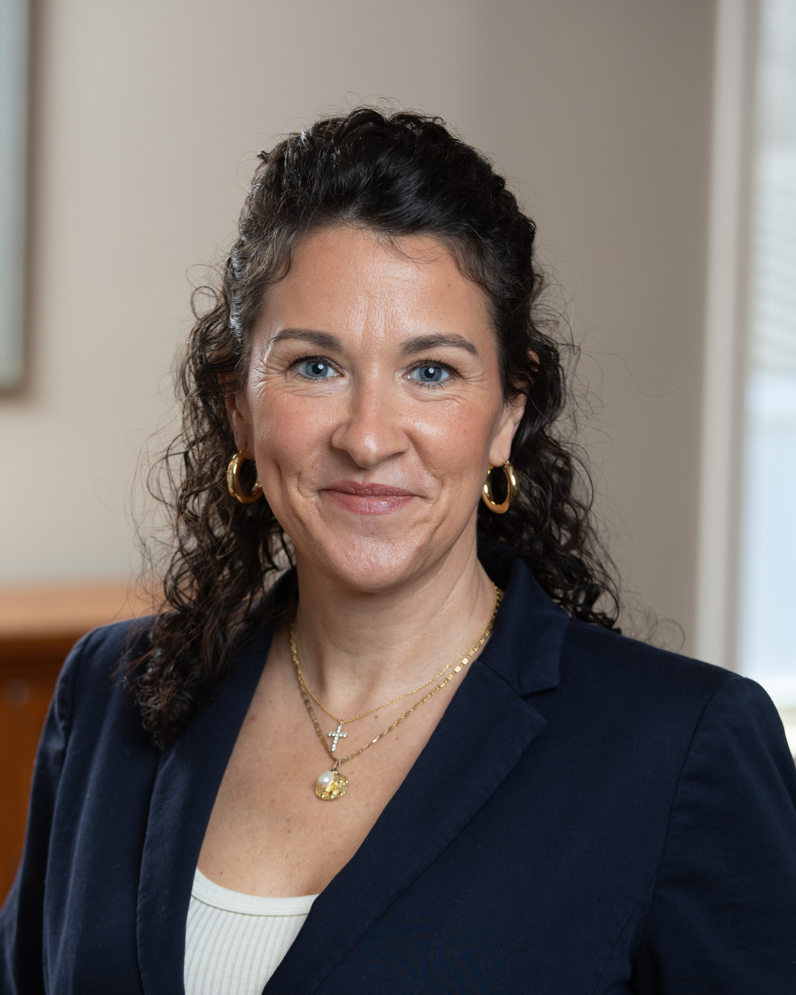 Elsa Giordano is a middle-aged woman with dark curly hair, blue eyes, wearing gold hoop earrings, layered necklaces, and a navy blazer, smiling indoors with a neutral background.