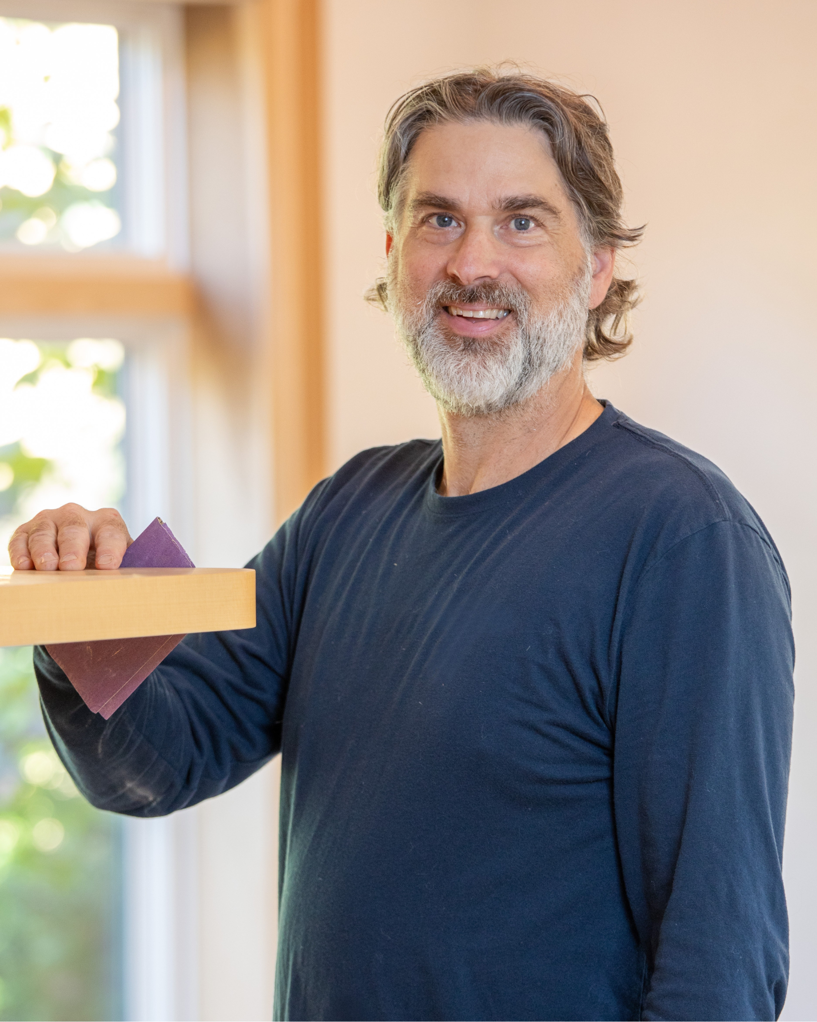 Sean Brines is a smiling middle-aged man with gray hair and beard, wearing a navy blue long sleeve shirt, sanding a wooden surface with sandpaper in a well-lit room.