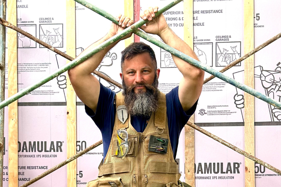 A man with a beard holds onto metal scaffolding with both hands at a construction site, pausing to look up at the camera. He is wearing a tan work vest with tools and safety glasses attached.