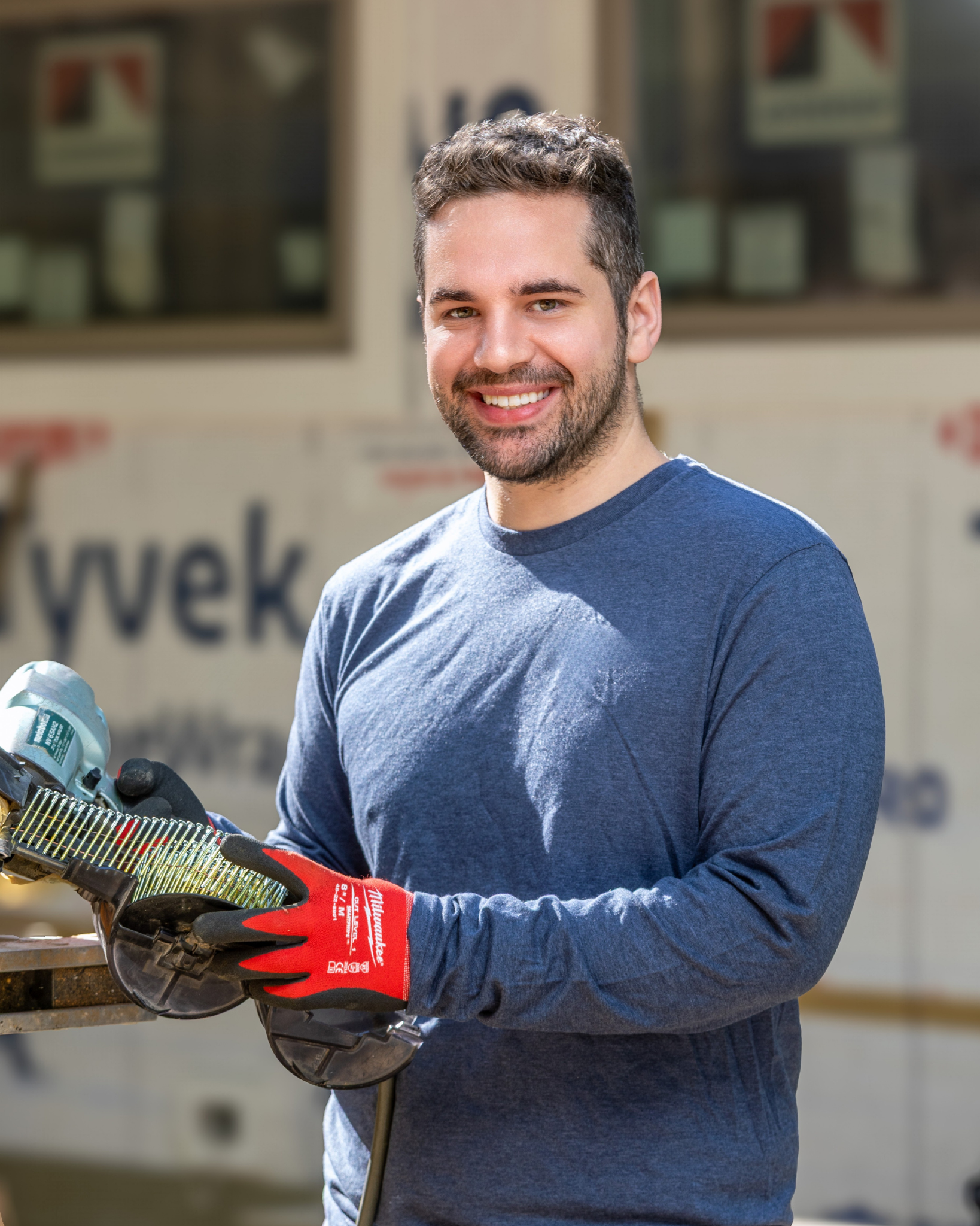 Deke Ludwig is a young man with short dark hair and a beard, wearing a long-sleeved blue shirt, smiling and holding a power saw while standing outdoors in front of a construction site.