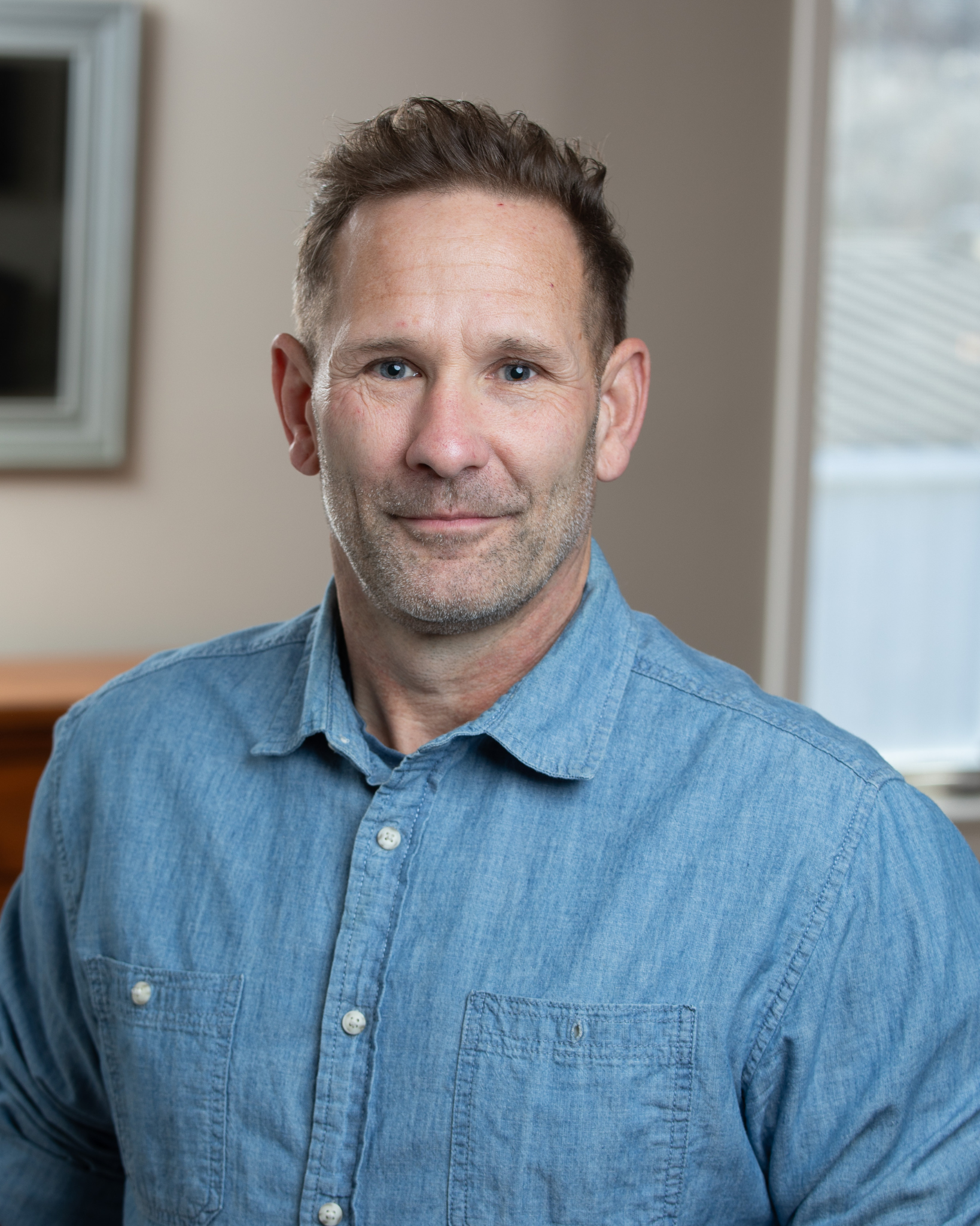 Abel Thomason is a middle-aged man with short brown hair and blue eyes, wearing a light blue denim shirt, sitting indoors near a window with natural light and a blurred background.