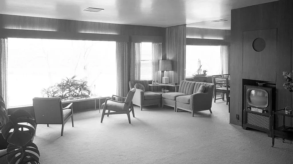 A black and white photo of a mid-century vintage living room with large windows, mid-century furniture, a television on a stand, and a small dining area in the background. This is a "Before" photo that shows a client's home before we renovated it.