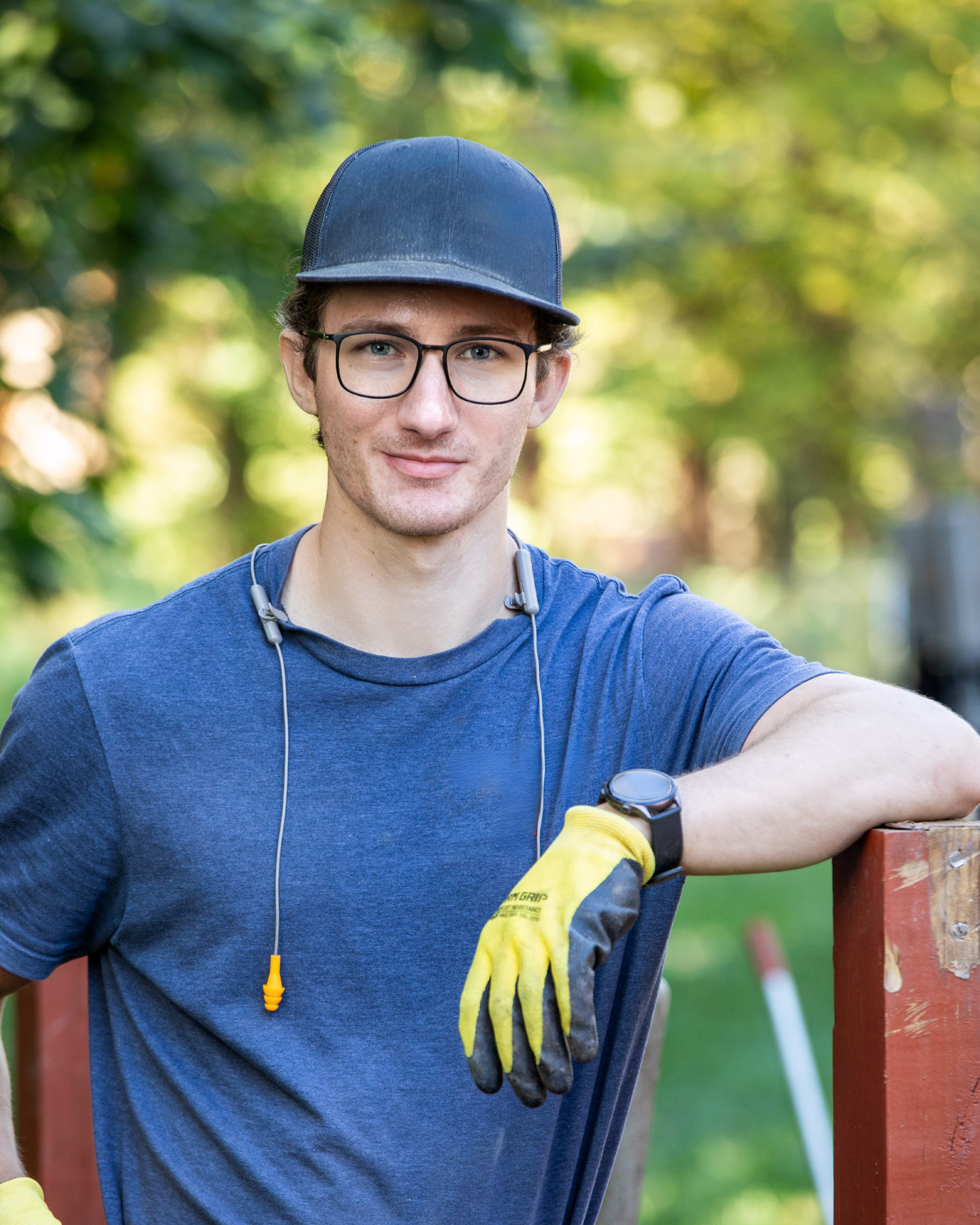 Gabriel Redente is a young man wearing glasses, a black cap, and a blue t-shirt, standing outdoors with a blurred green background. He has yellow gloves on and a smartwatch on his left wrist, with earplugs around his neck, leaning on a railing.