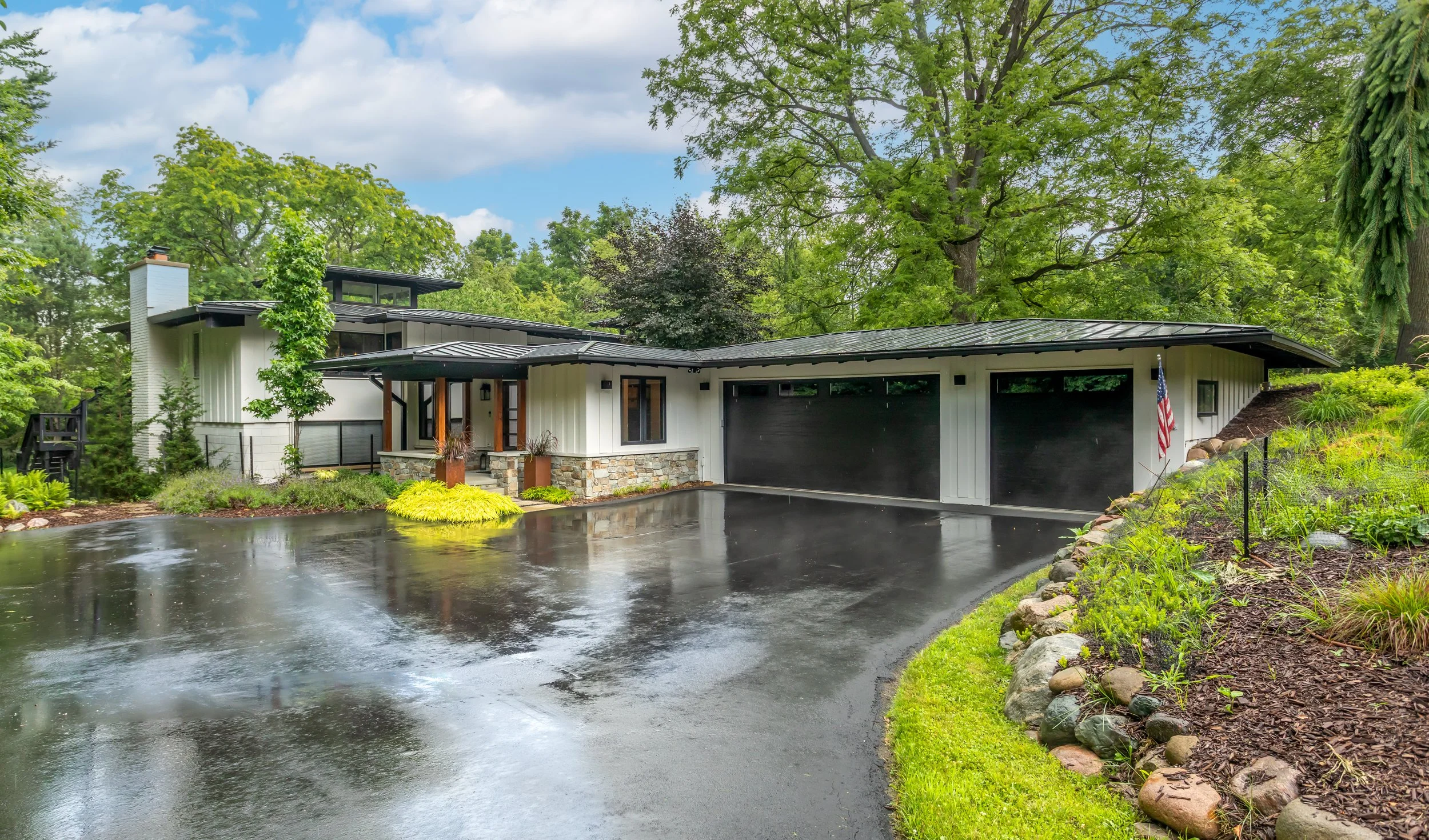 Modern custom luxury home with white exterior, black garage doors, surrounded by green trees and landscaping, with wet driveway.
