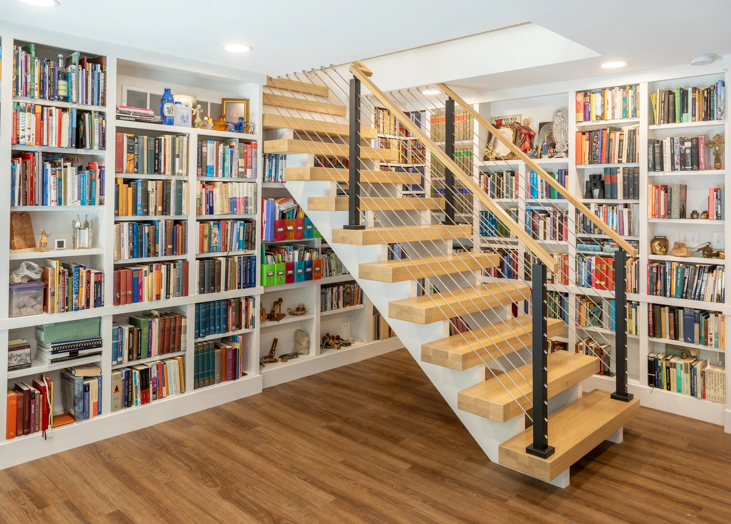 A spacious home library with white bookshelves filled with colorful books, a modern open wooden staircase with metal railings, and hardwood floors.