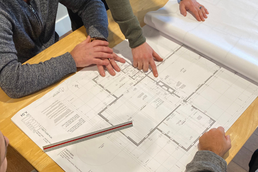 Three people examining large architectural blueprints showing a floor plan of a custom home, spread out on a wooden table.