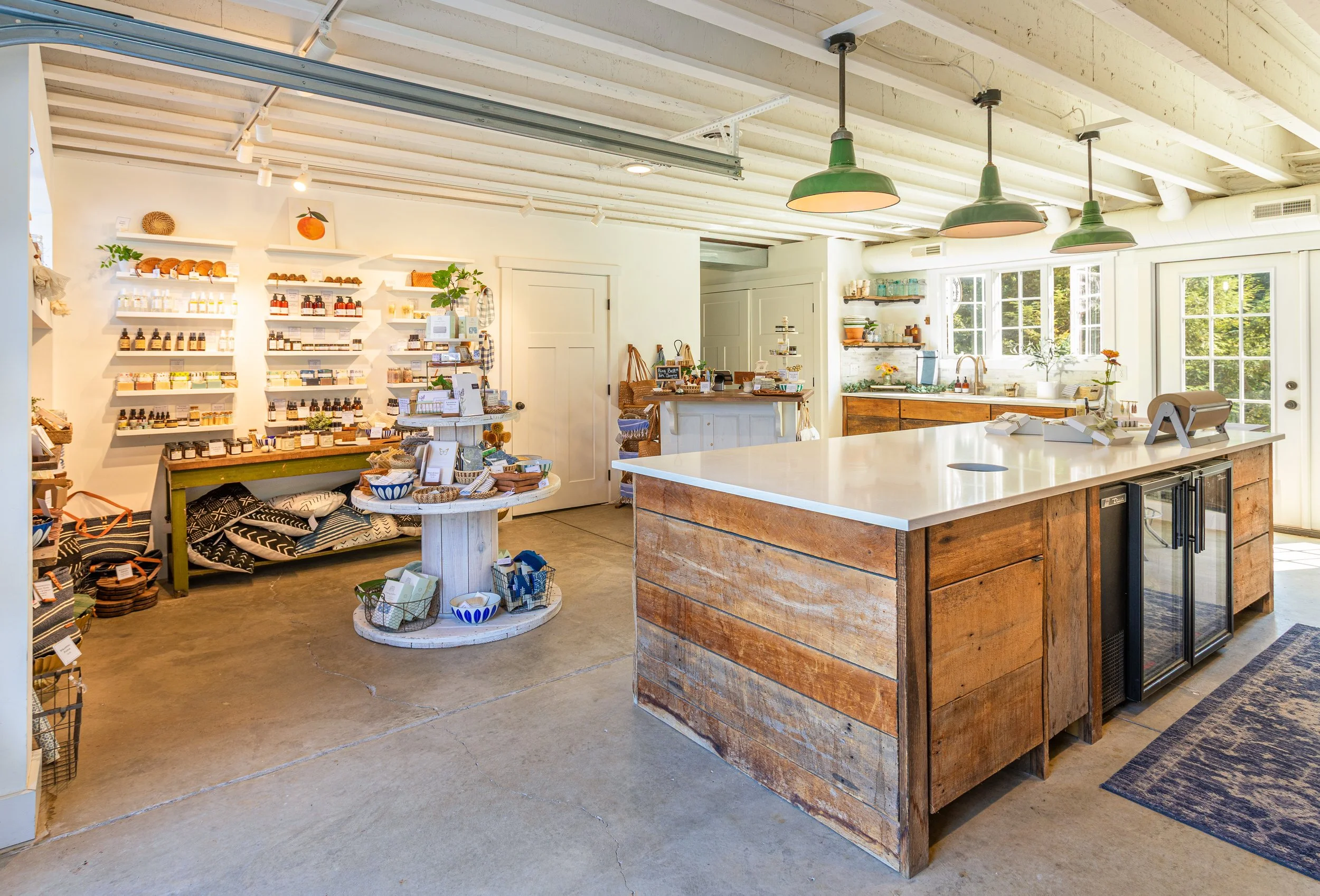 Interior of a cozy shop called Little Flower Soap Co., with white walls and ceiling, natural wood accents, green pendant lights, large windows, a kitchen counter, and shelves with various products on display.