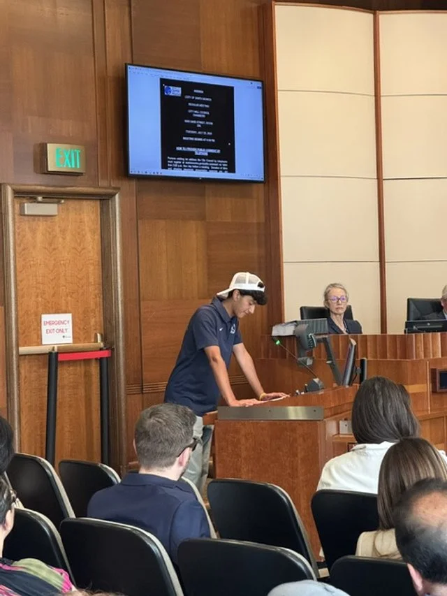 A person wearing a Virtual Reality headset standing at a podium in a conference room with an audience, while two women are seated behind a desk at the front of the room.