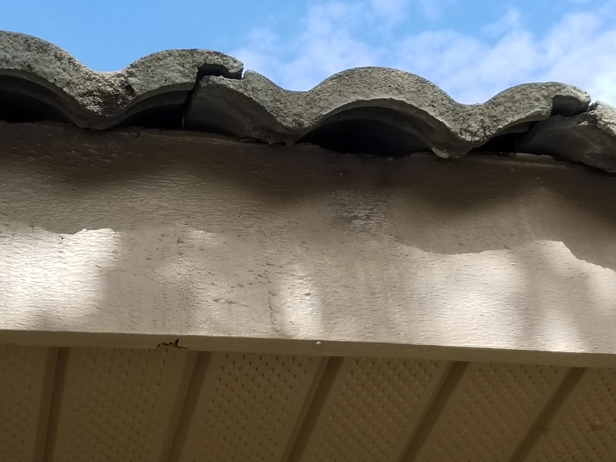 Close-up of a roof edge with weathered gravel tiles and a section of a beige soffit underside, with a blue sky and clouds in the background.
