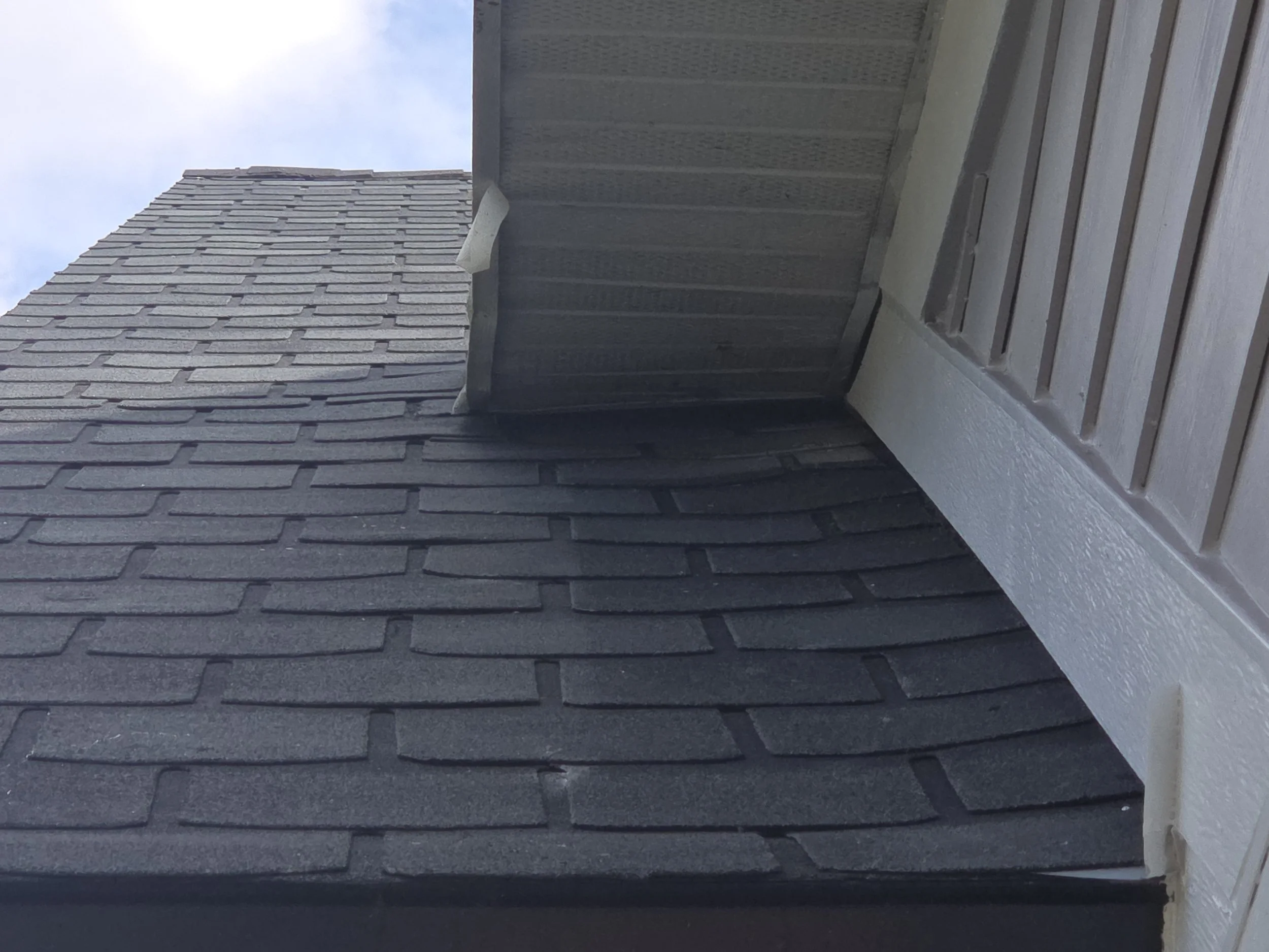 Close-up view of a roof with dark gray asphalt shingles, showing the corner of the roof and part of the exterior wall with beige siding and white trim.