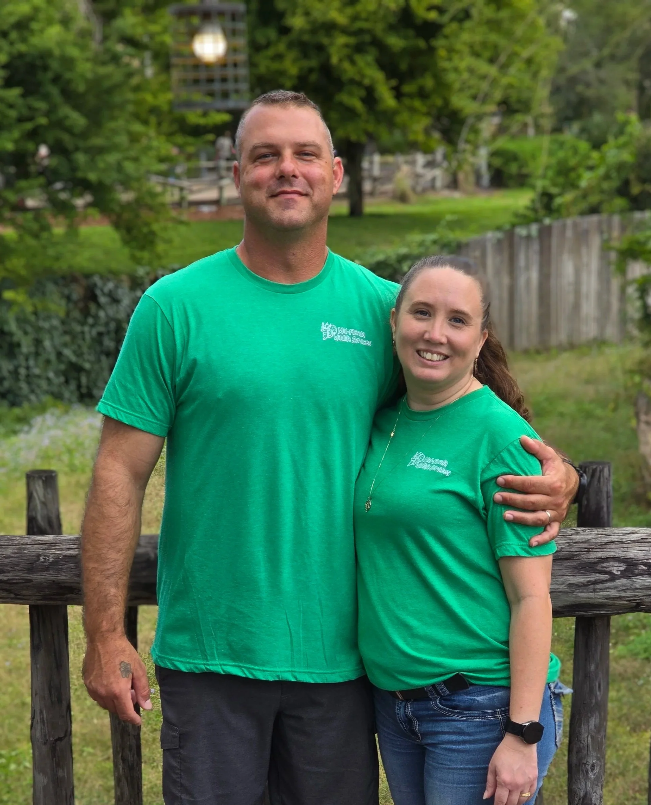 A man and a woman standing outdoors together, both wearing matching green T-shirts, smiling and embracing each other. The background features green trees, a fence, and a wooden railing.