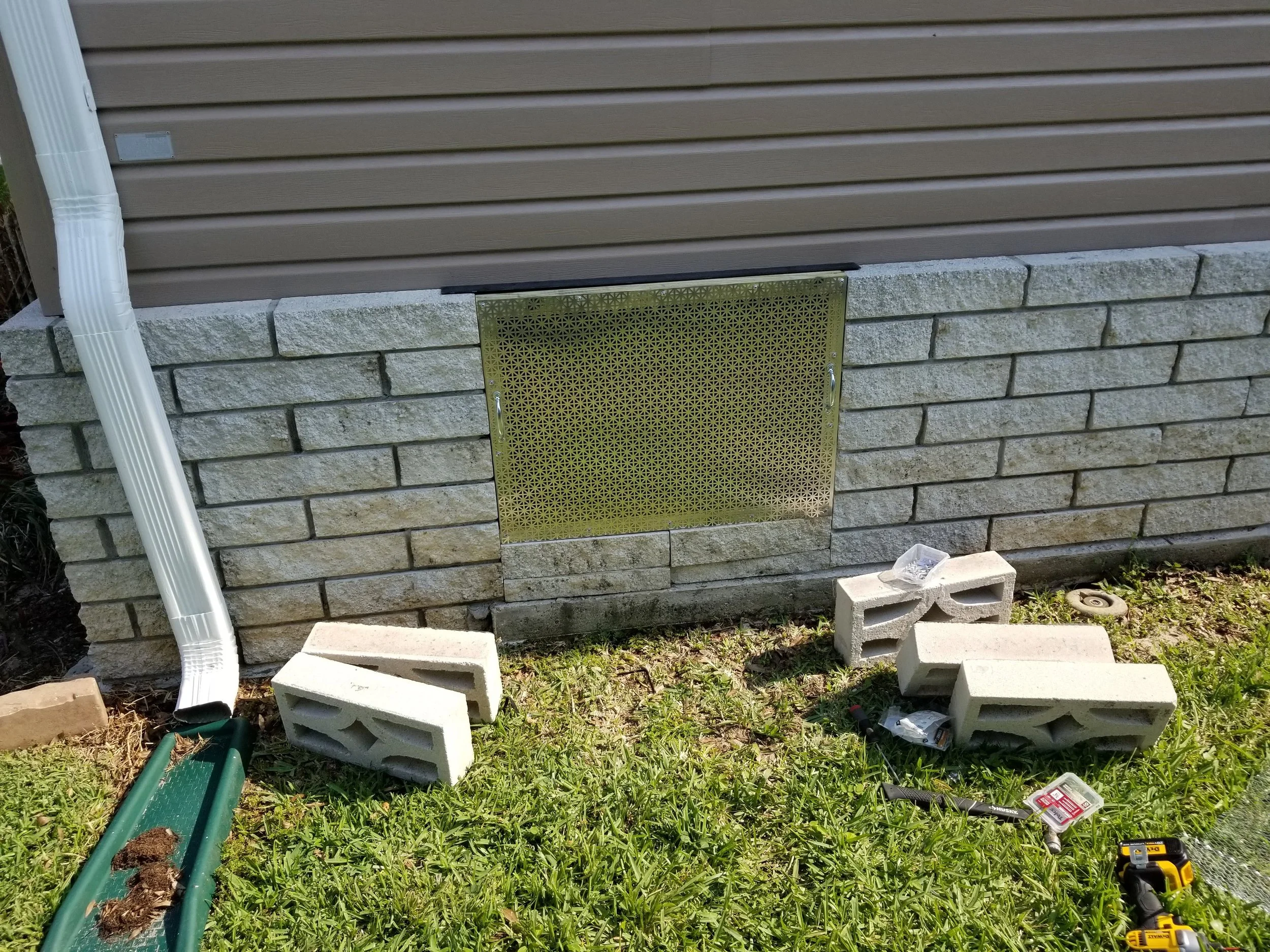 Construction work in progress outside a house, with bricks and tools on the ground, a wall vent, and a downspout on the side.