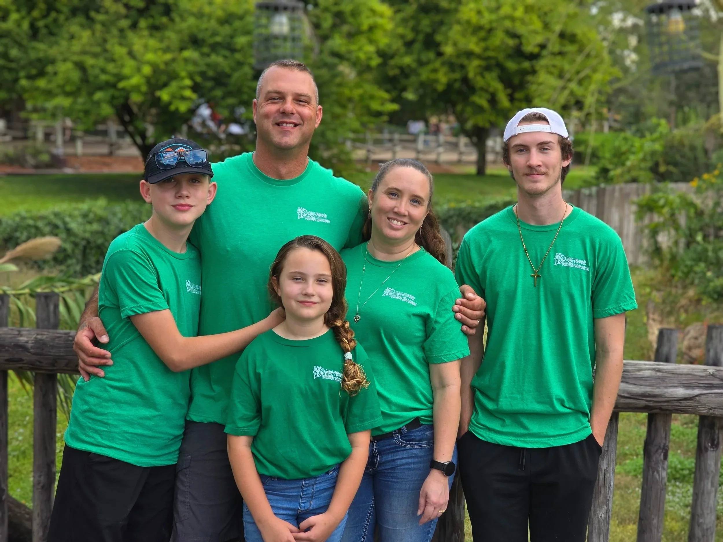 A family of five standing outdoors in front of a wooden fence with trees and greenery behind them. They are all wearing matching green T-shirts.