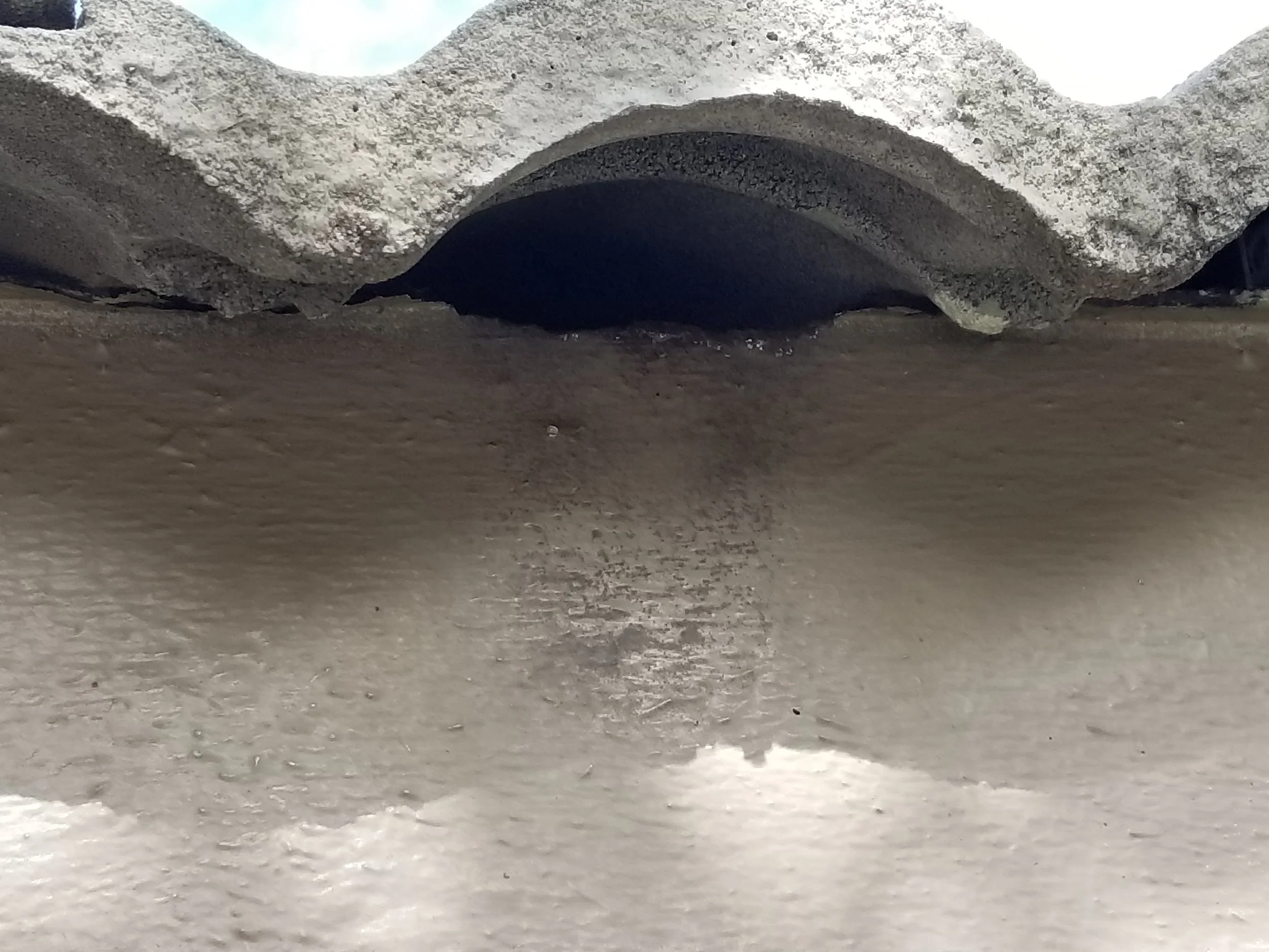 Close-up of a narrow drainage pipe opening beneath a concrete water feature with water flowing out.
