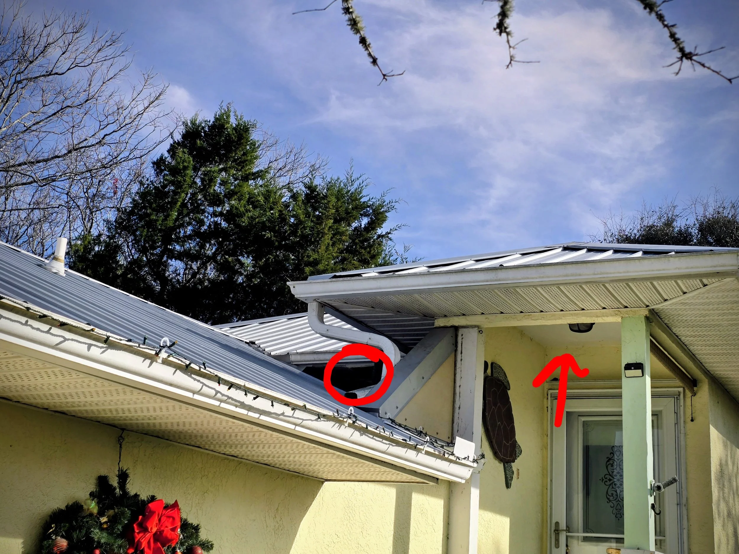 Close-up of a house roof with a white gutter system, a water drainage pipe, and a roof vent circled in red. There is also an arrow pointing upward toward the roof structure. The house is painted yellow and there is a decorated Christmas wreath below 