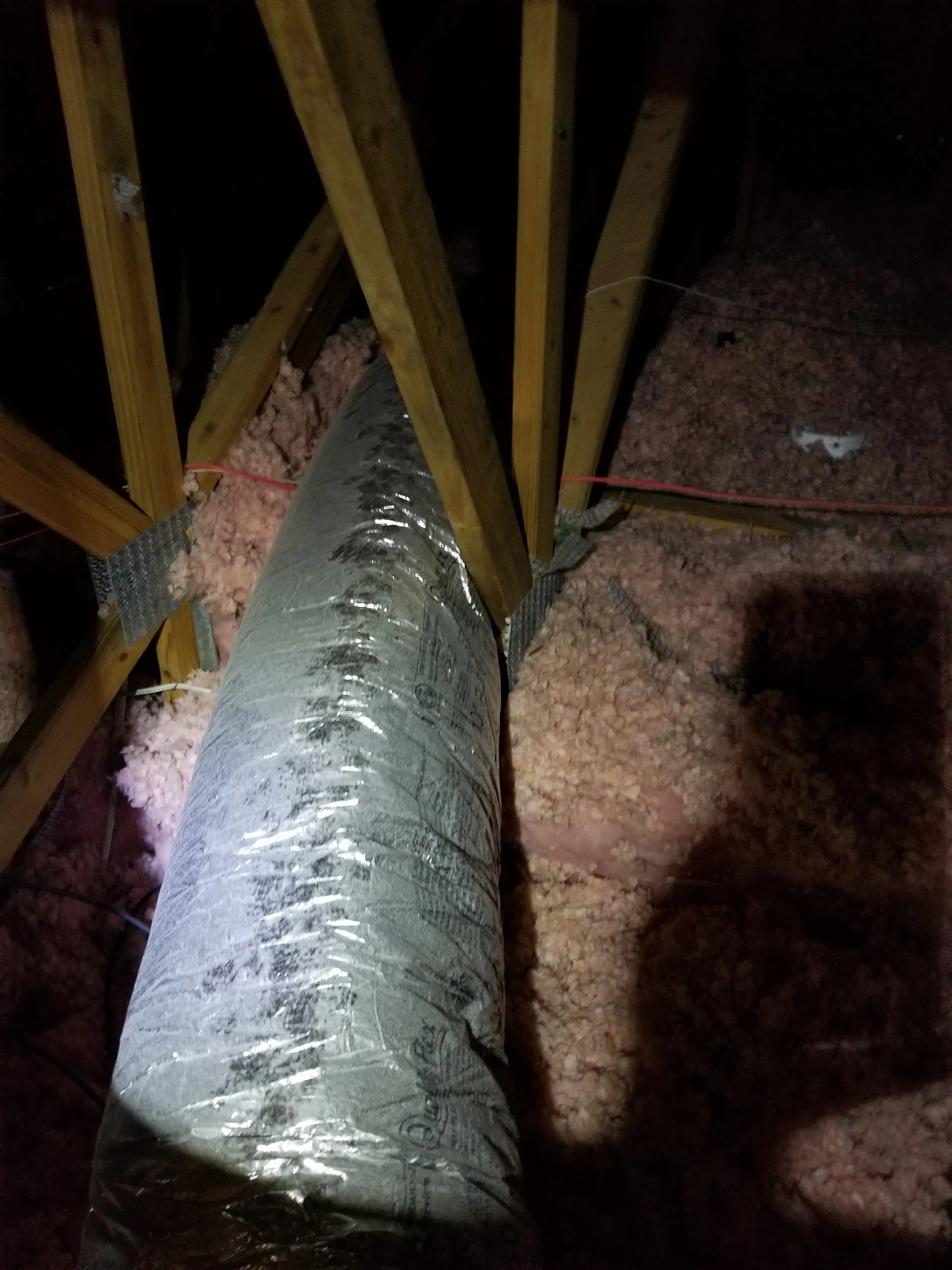 View of a construction space showing a large duct pipe, wooden framing, pink insulation, and electrical wiring.