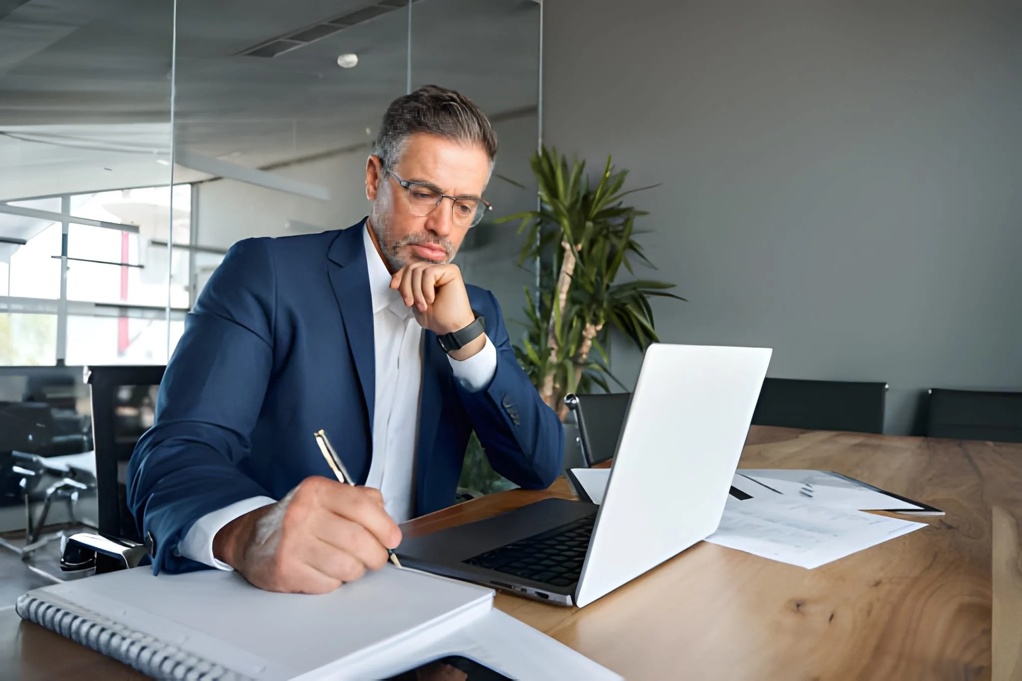 A businessman wearing a navy suit, white shirt, and glasses sitting at a wooden table in an office, writing on a notebook with a pen while looking at a laptop. There are documents on the table and a potted plant behind him.