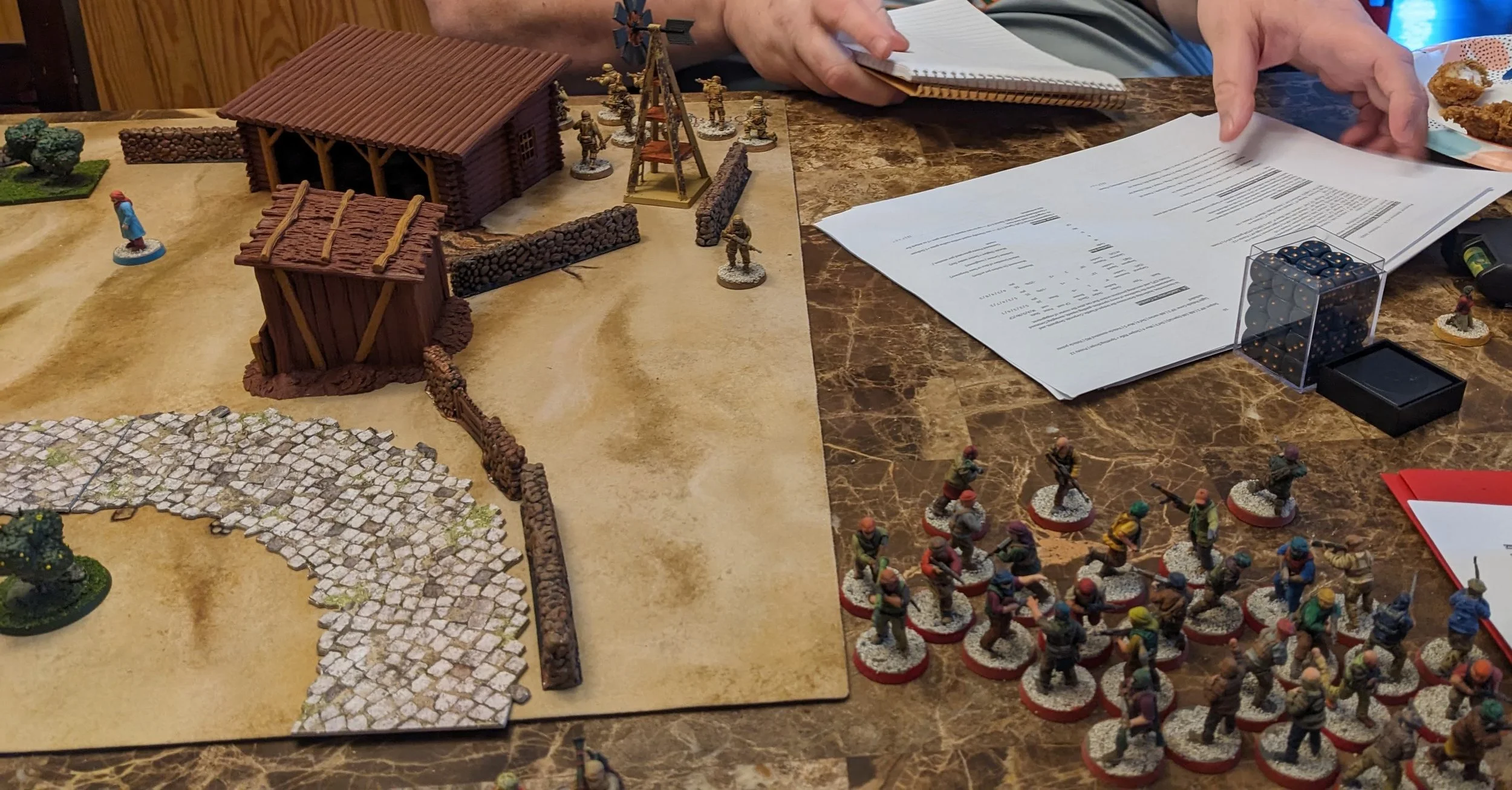 Miniature soldiers and buildings on a tabletop for a tabletop game, with dice and rule books nearby.