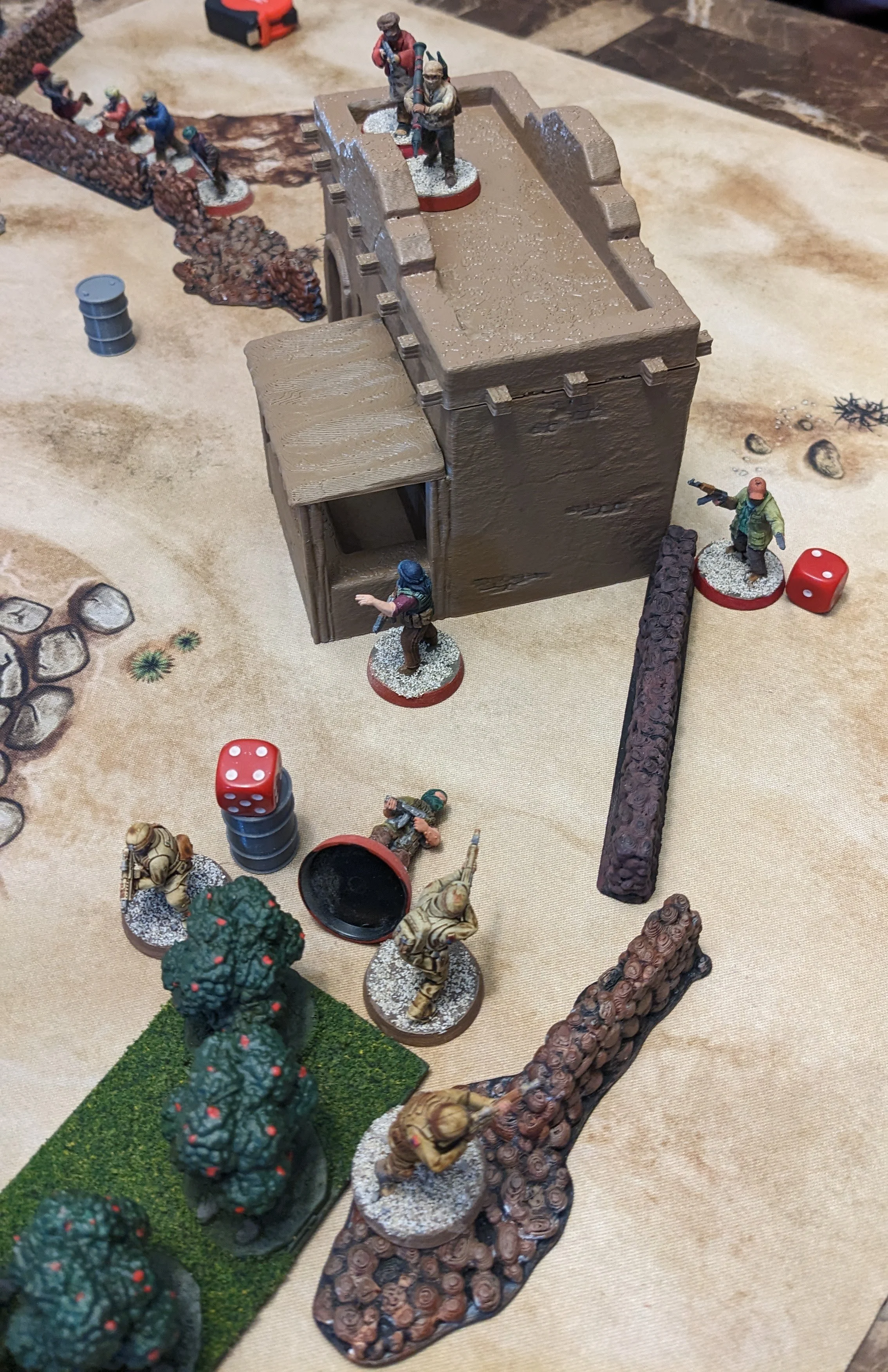 Tabletop war game with miniature soldiers, a small tower, trees, and dice on a desert-themed game mat.