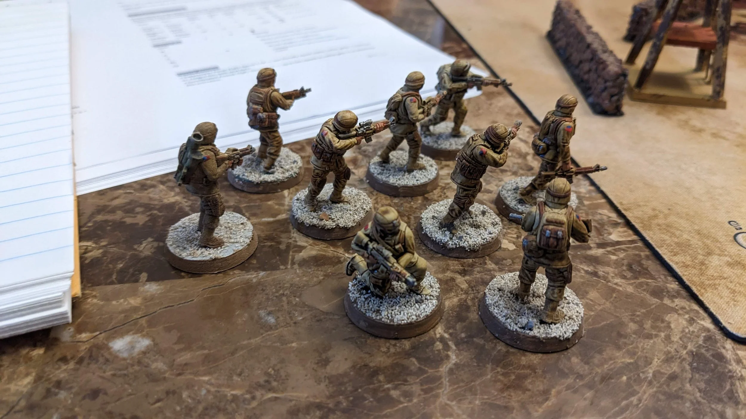 Set of miniature soldier figurines painted in military style, arranged on a tabletop with scattered sand and small props, suggesting a diorama or gaming scene.