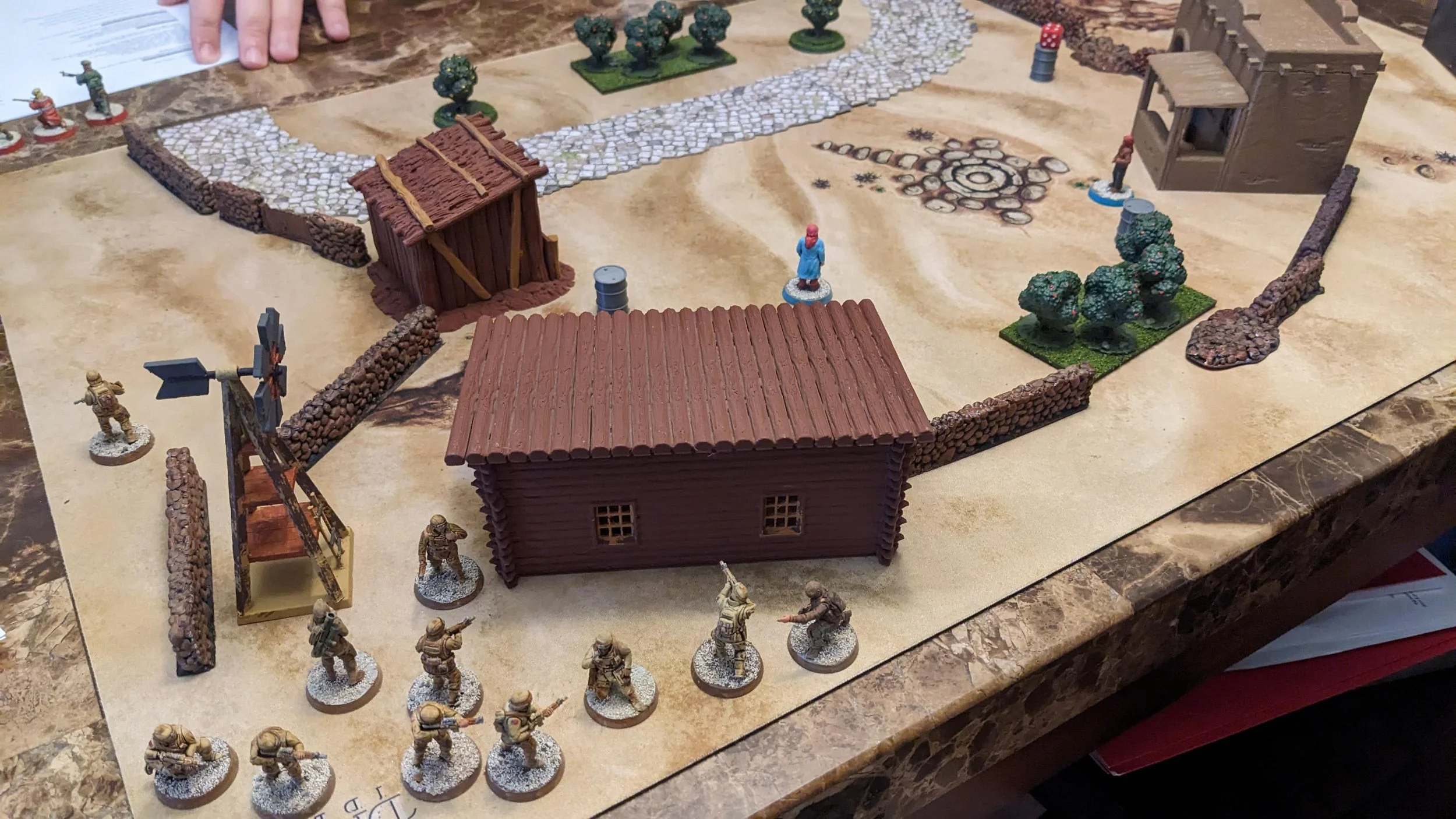 Tabletop wargame setup with miniature soldiers, buildings, trees, rocks, and game tokens on a mat designed to look like a dirt battlefield.