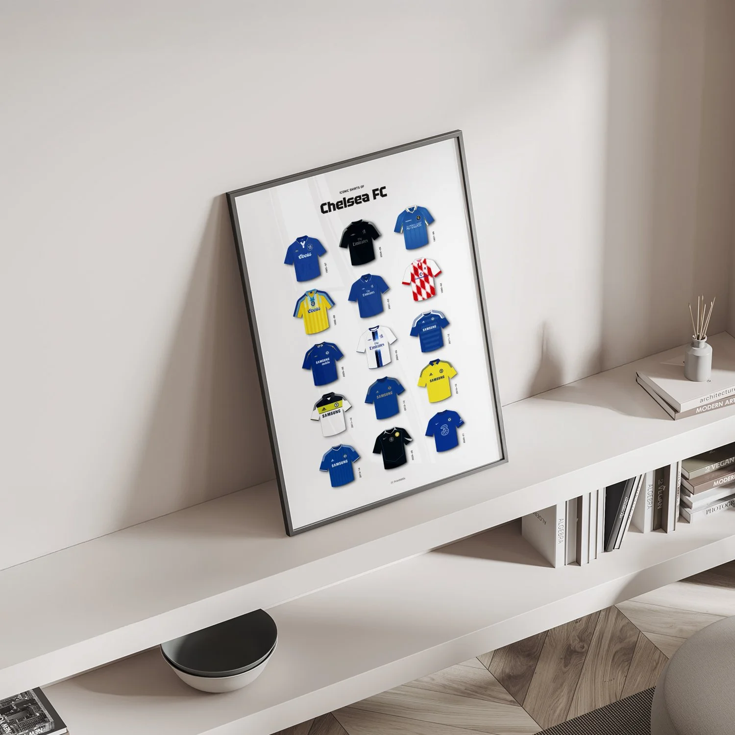 Framed Chelsea football kit evolution poster leaning on a modern white shelf with books and decor.