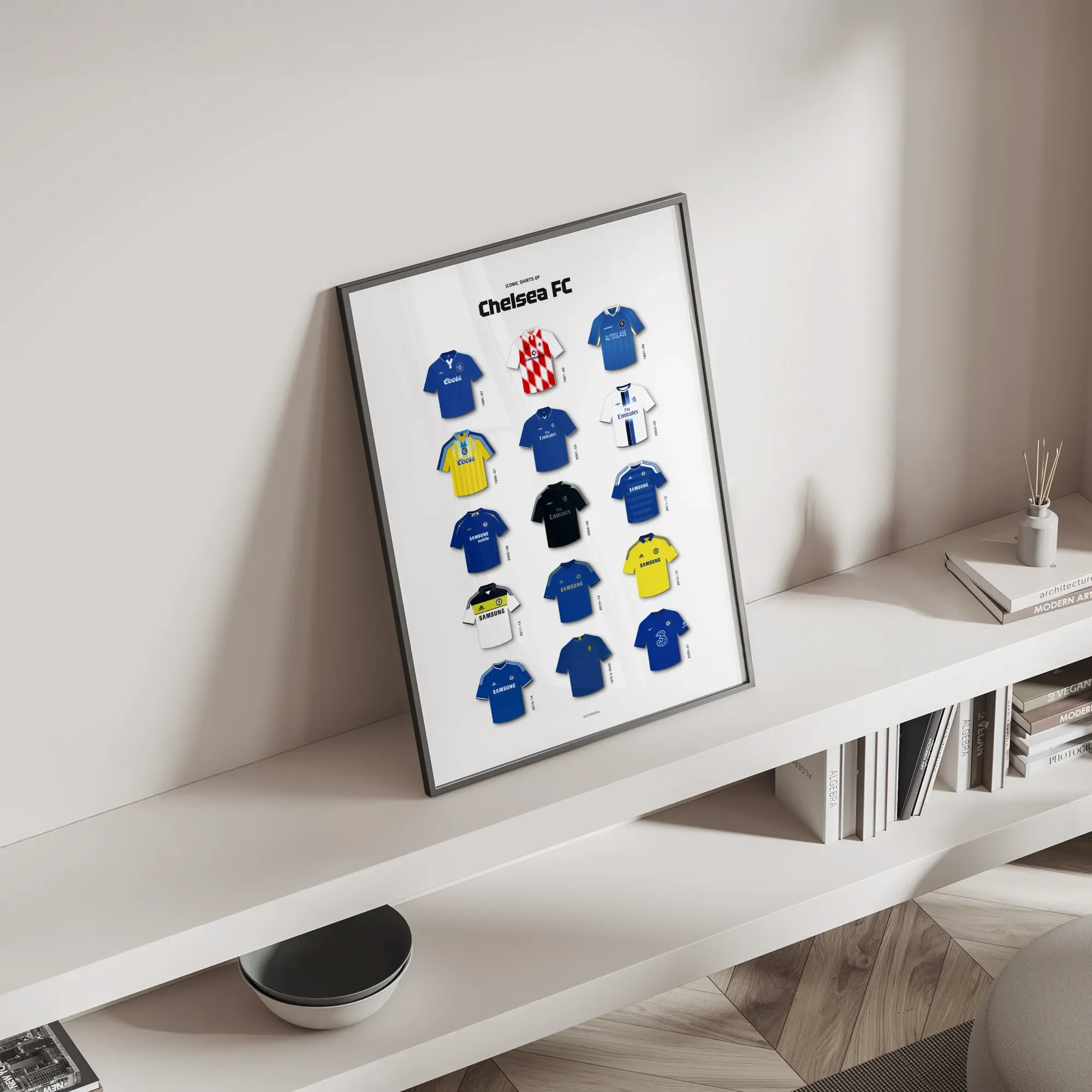 Framed Chelsea football shirt poster leaning on a minimalist white shelf in a bright room.