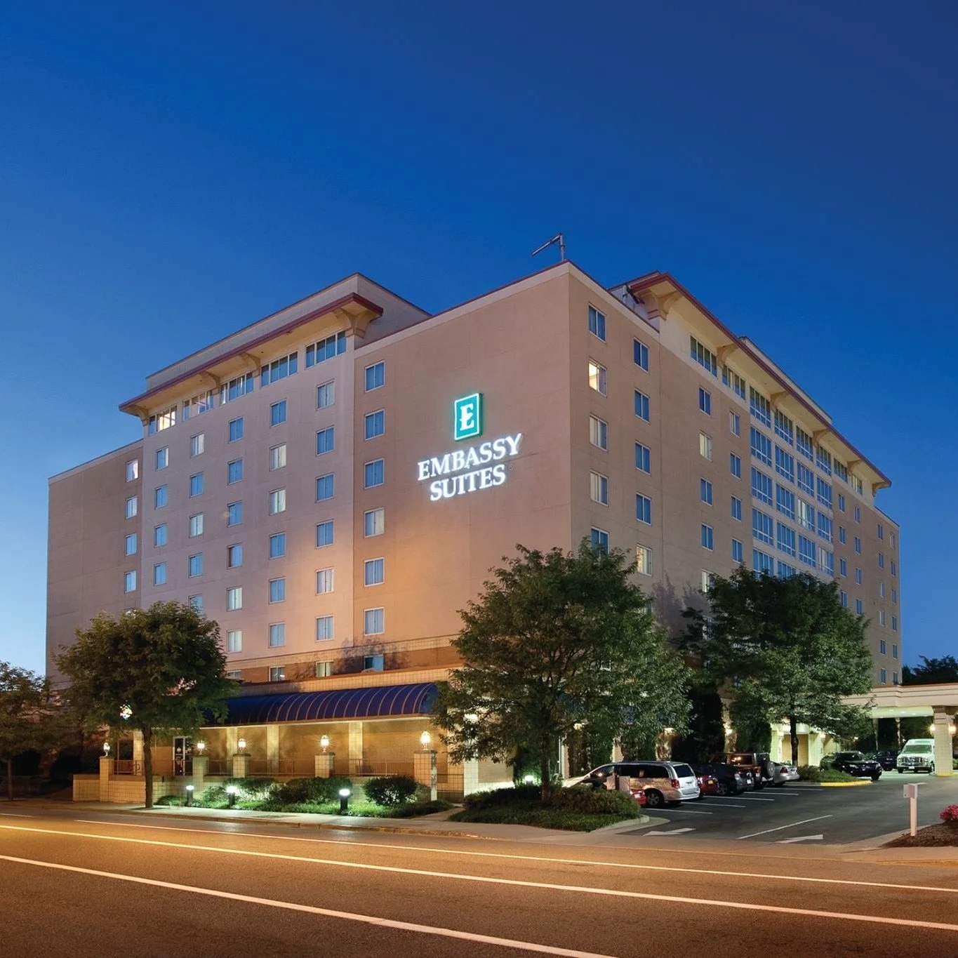 Nighttime exterior view of a tall hotel building labeled 'Embassy Suites' with illuminated sign, trees, parked cars, and a well-lit entrance.
