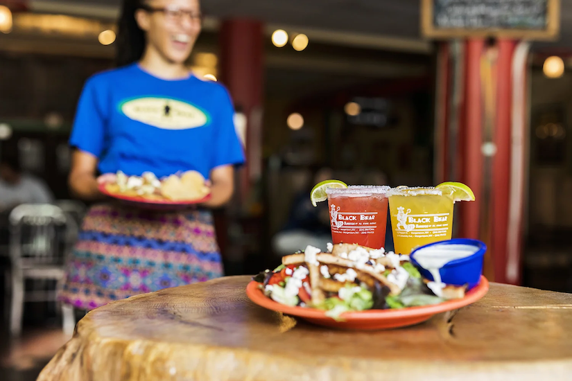A wooden table with a plate of salad, two cups of soda, and a small bowl of sauce in a restaurant setting. A smiling waitress in a blue shirt with a name tag and colorful skirt stands in the background.