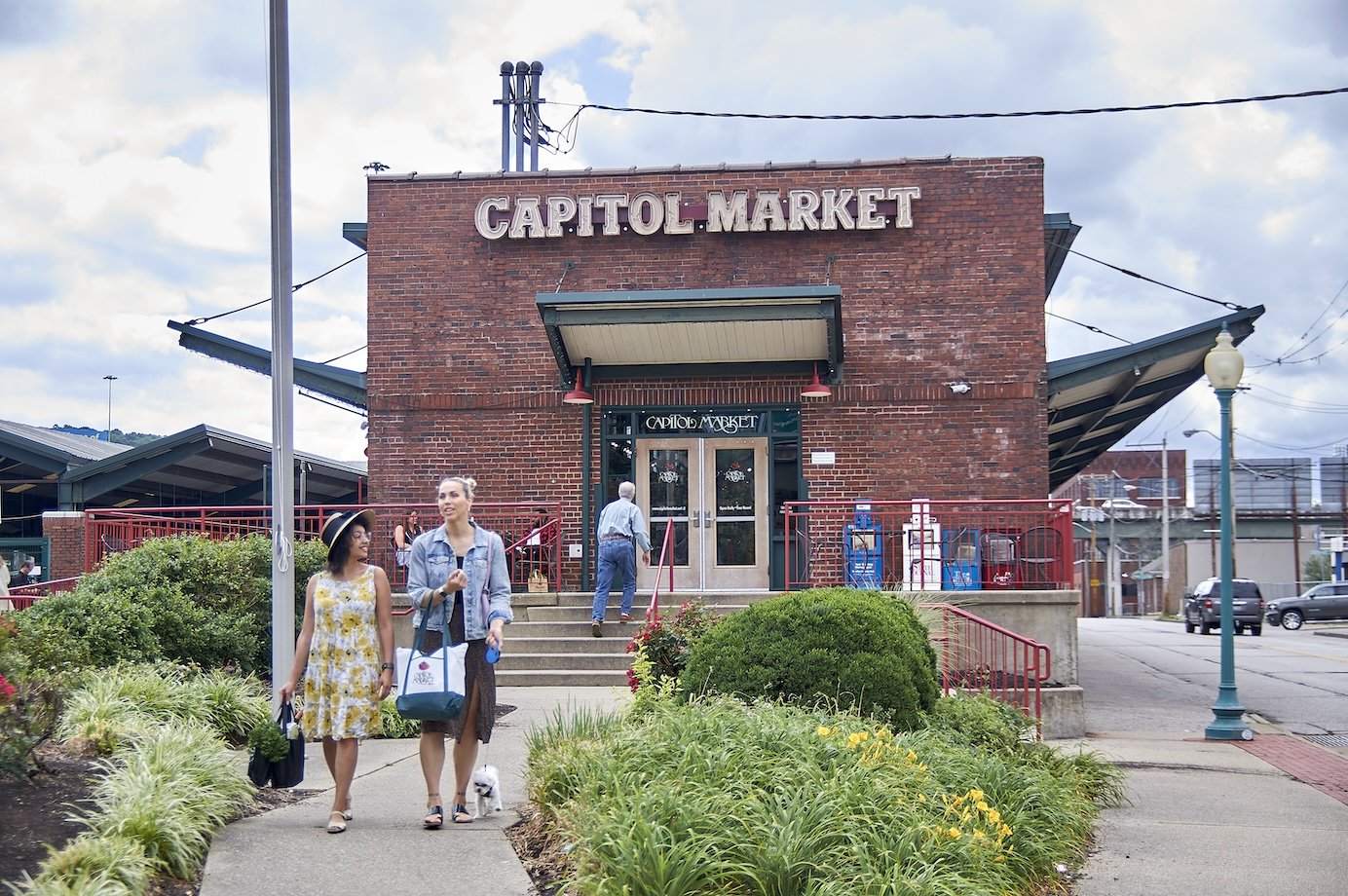 Exterior view of Capitol Market building with people walking in front, greenery, and parking lot.