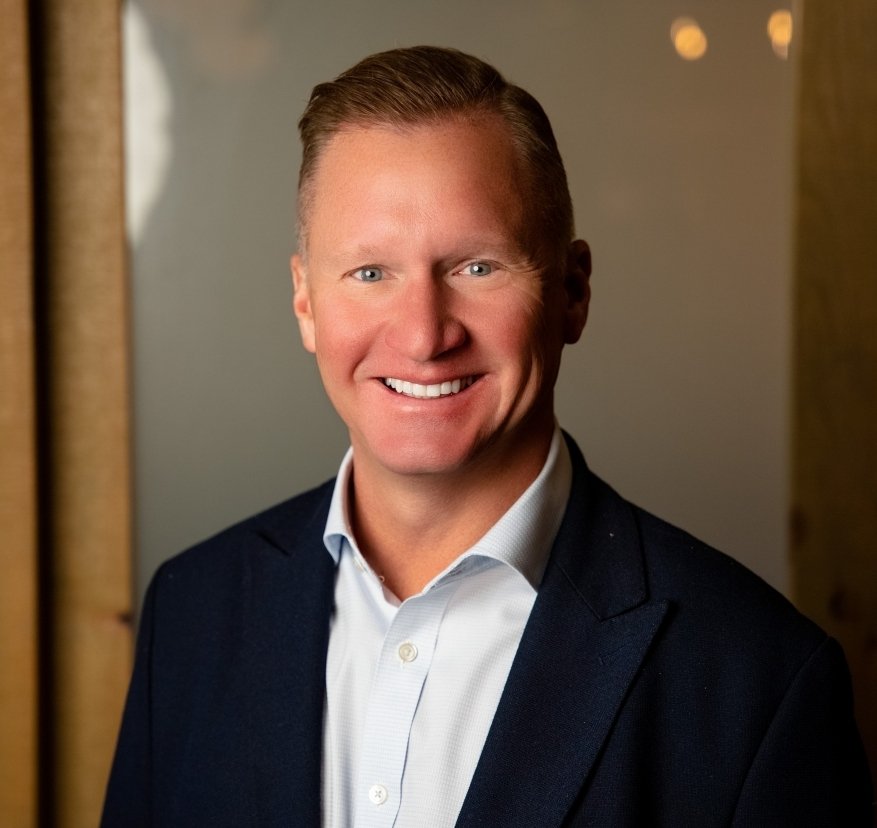 Smiling man in a suit and white shirt with wooden background and warm lighting.