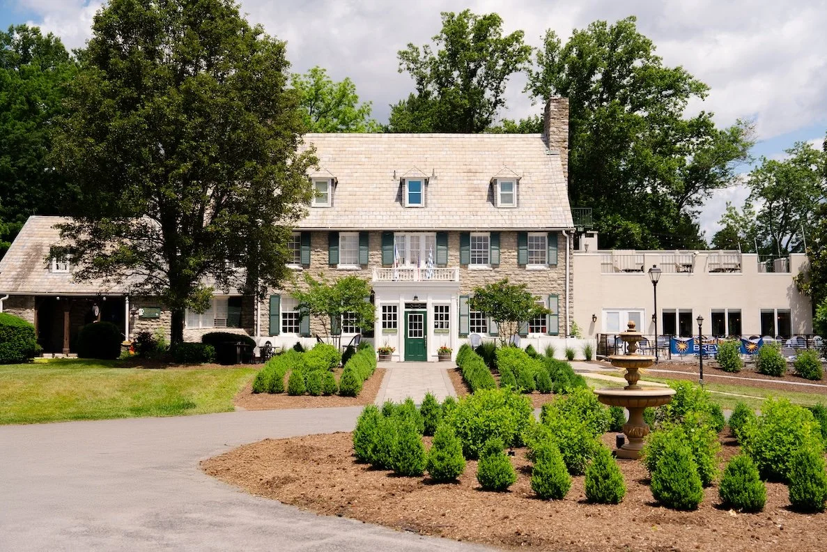 Large historic stone house with a steep roof and multiple windows, surrounded by landscaped gardens, trees, and a paved walkway, under cloudy skies.
