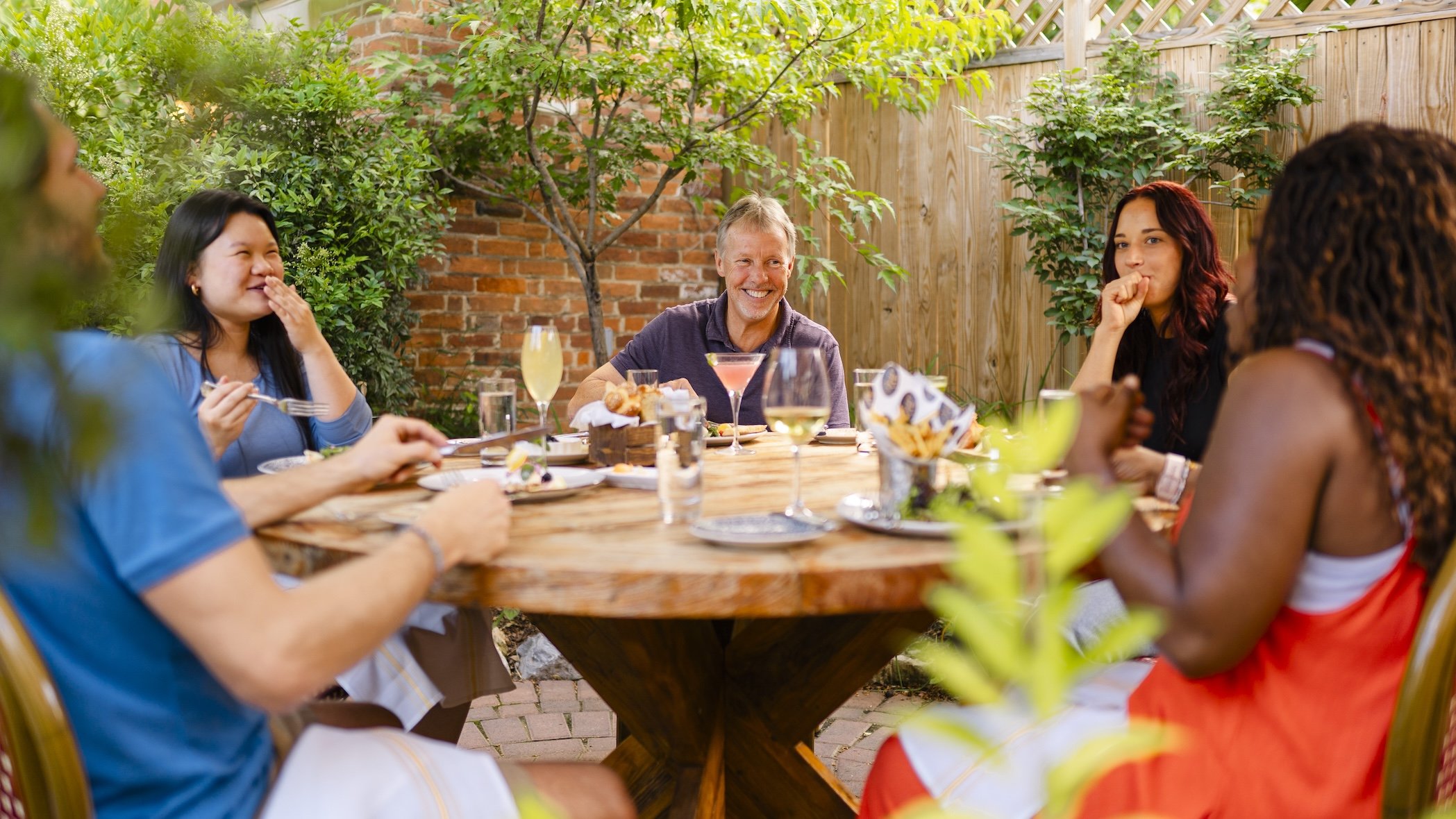 Group of six friends sitting at outdoor wooden table, enjoying a meal and drinks in a backyard with trees and a wooden fence, smiling and engaging in conversation.