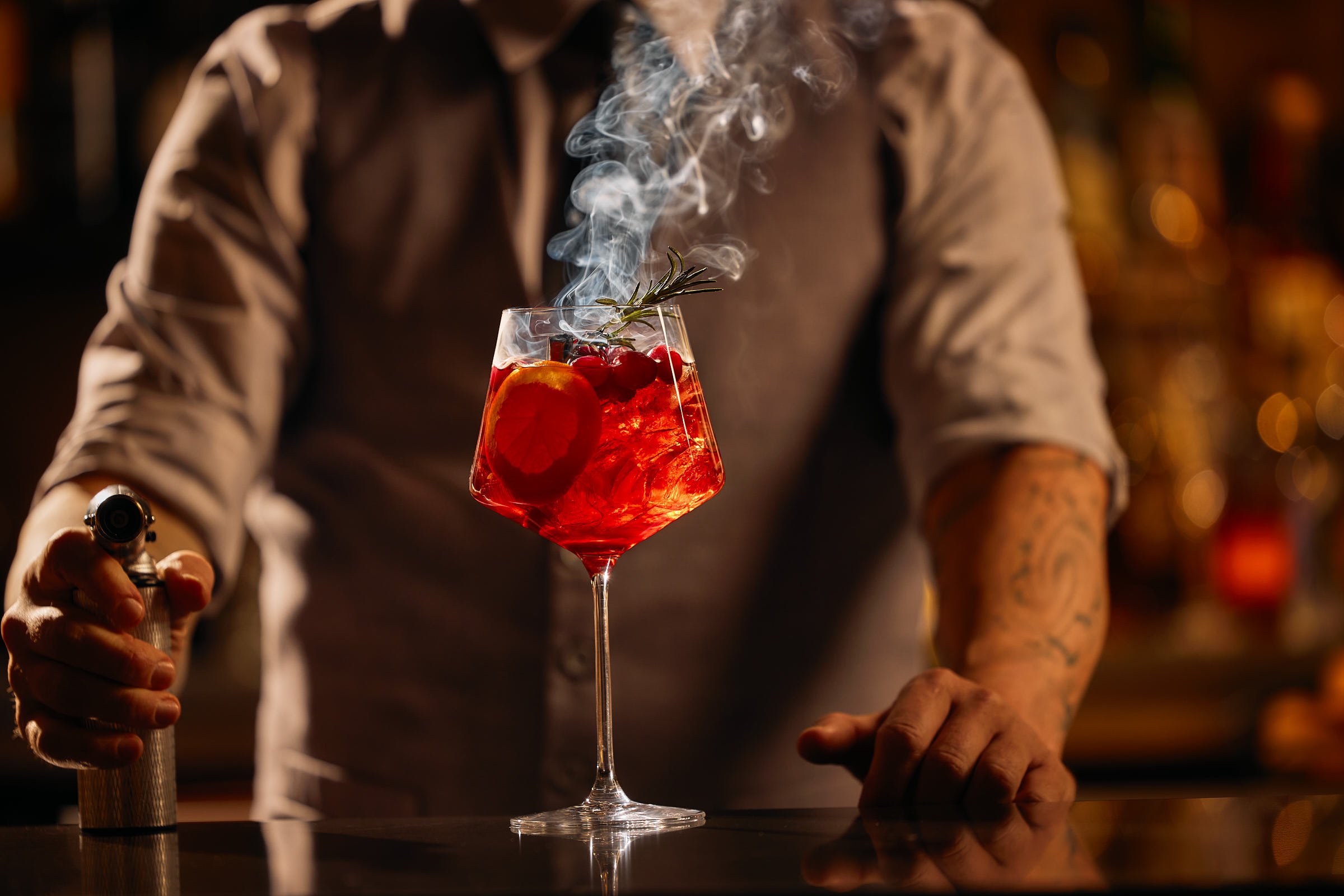 A bartender adds smoke to a vibrant red cocktail garnished with fruit and herbs, held by a long-stemmed glass.