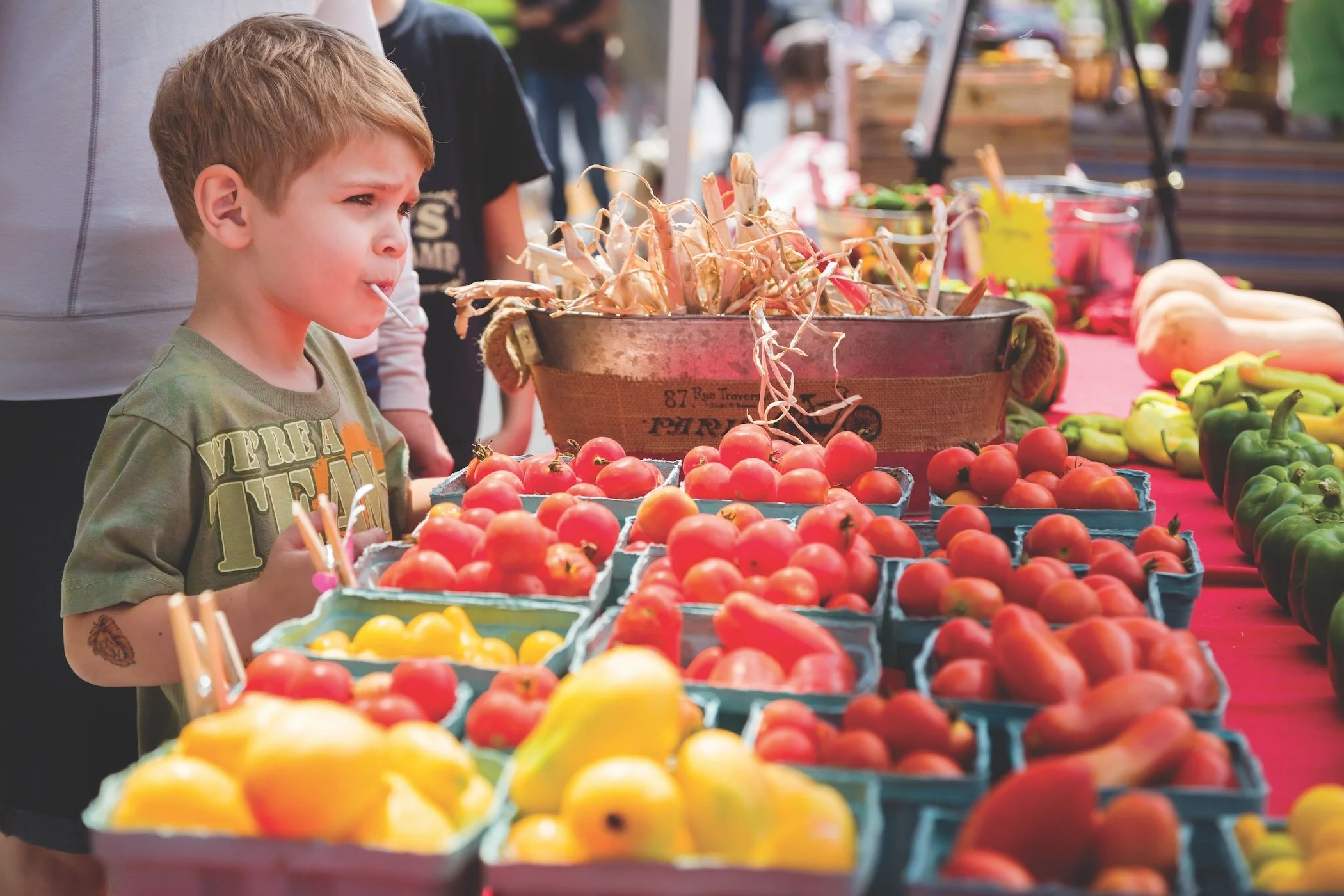 A young boy at a farmers market examining a display of cherry tomatoes with a lollipop in his mouth. The table has various fresh vegetables and fruits like yellow and red tomatoes, bell peppers, and gourds, with more produce and market stalls in the background.