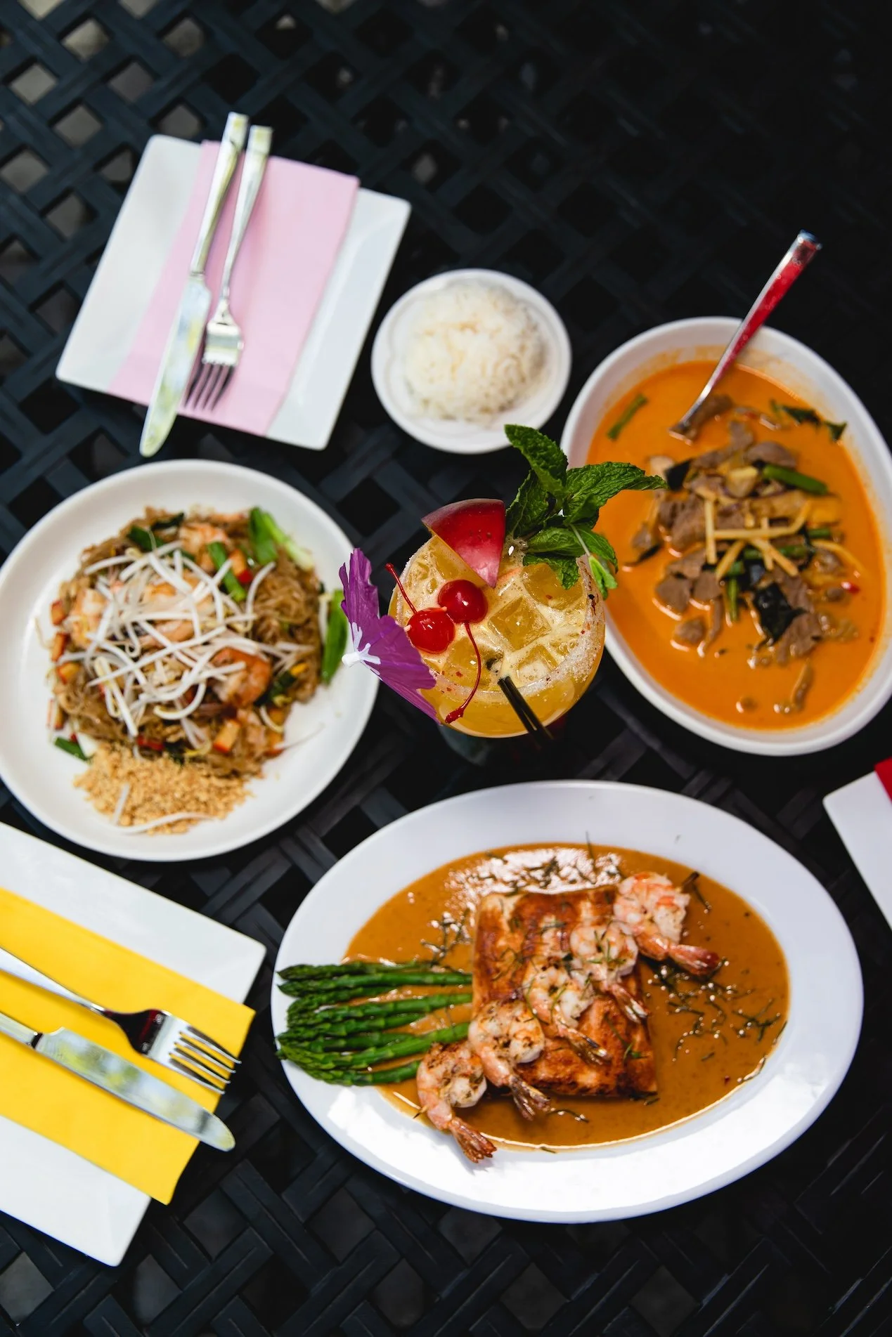 An assortment of Thai dishes on a black table, including a bowl of red curry with meat and vegetables, a green vegetable dish with rice, a plate of fried rice with seafood, and a plate of grilled shrimp with asparagus, alongside a colorful cocktail garnished with fruit and mint.