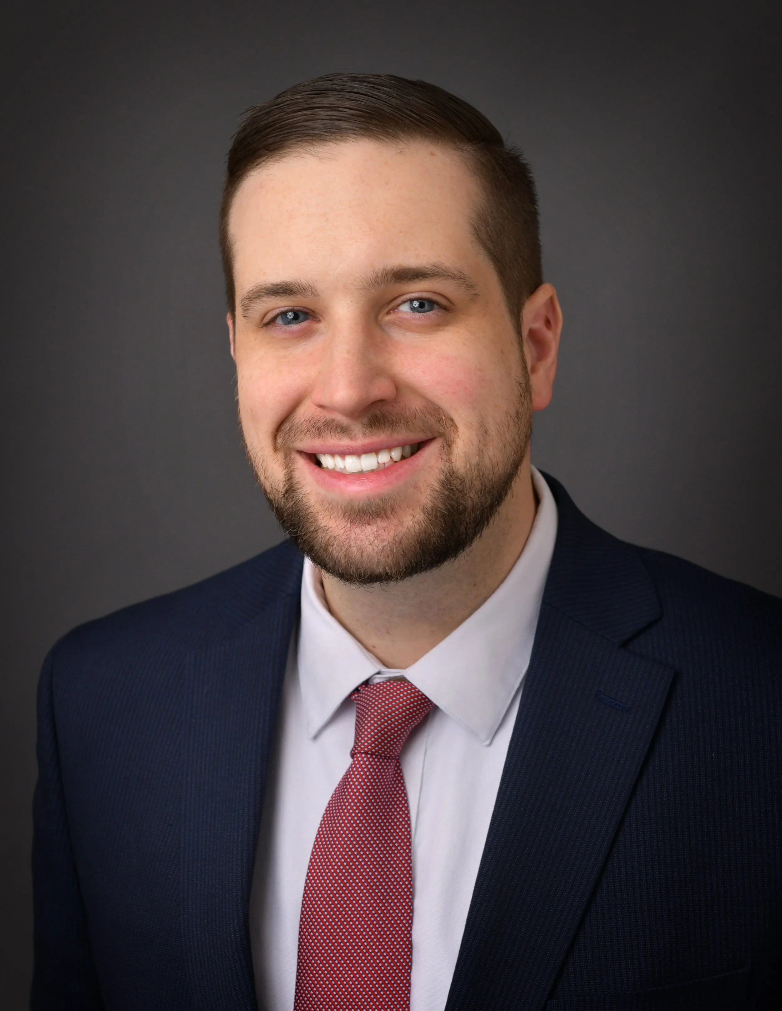 Professional headshot of a young man with brown hair and a beard, wearing a dark suit, white shirt, and red tie, smiling against a dark background.