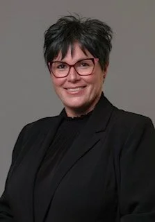 A woman with short dark hair, wearing glasses and a black blazer, smiling against a gray background.