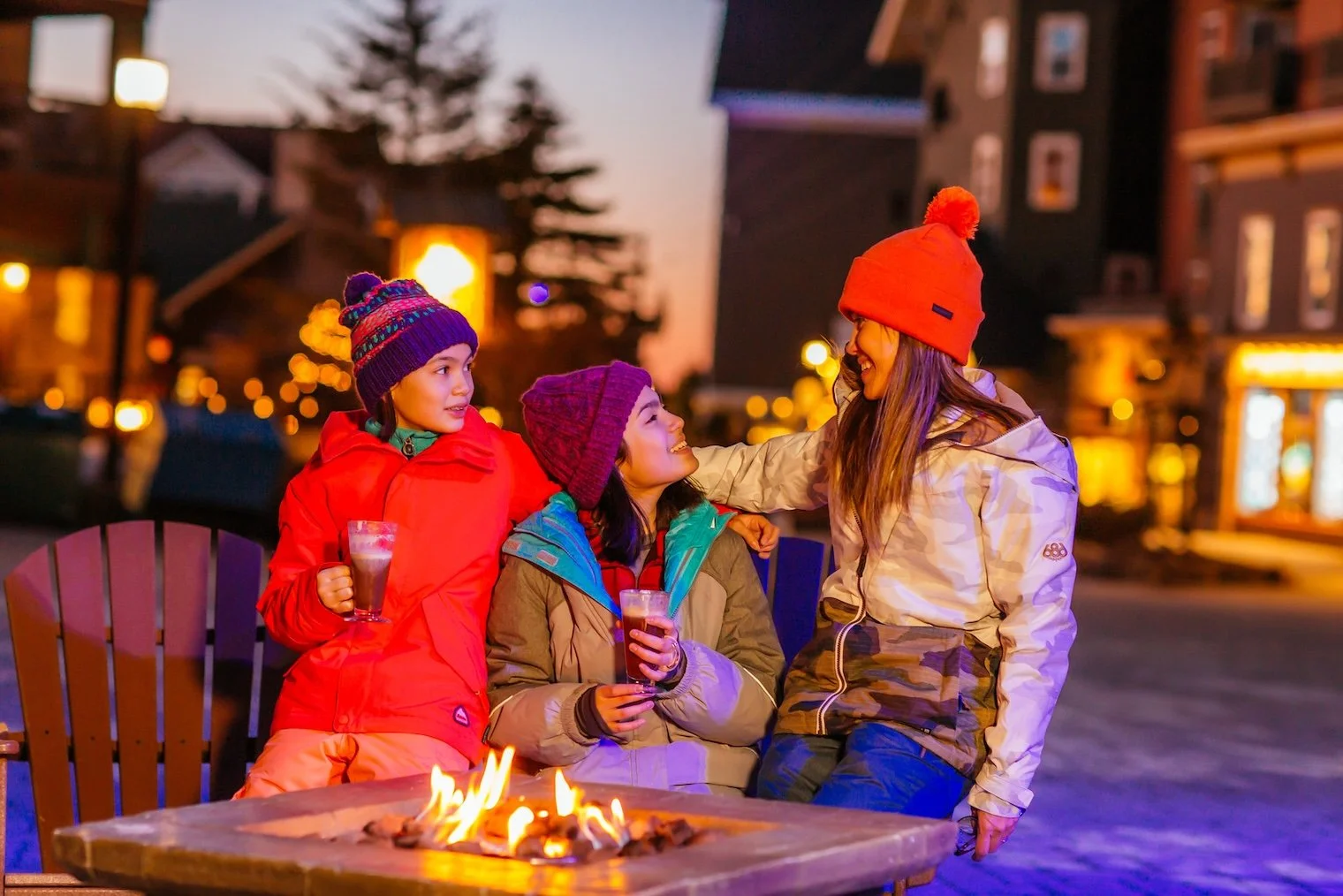 A group of three women enjoying beers around a fire pit outdoors during dusk, wearing colorful winter jackets and beanies.